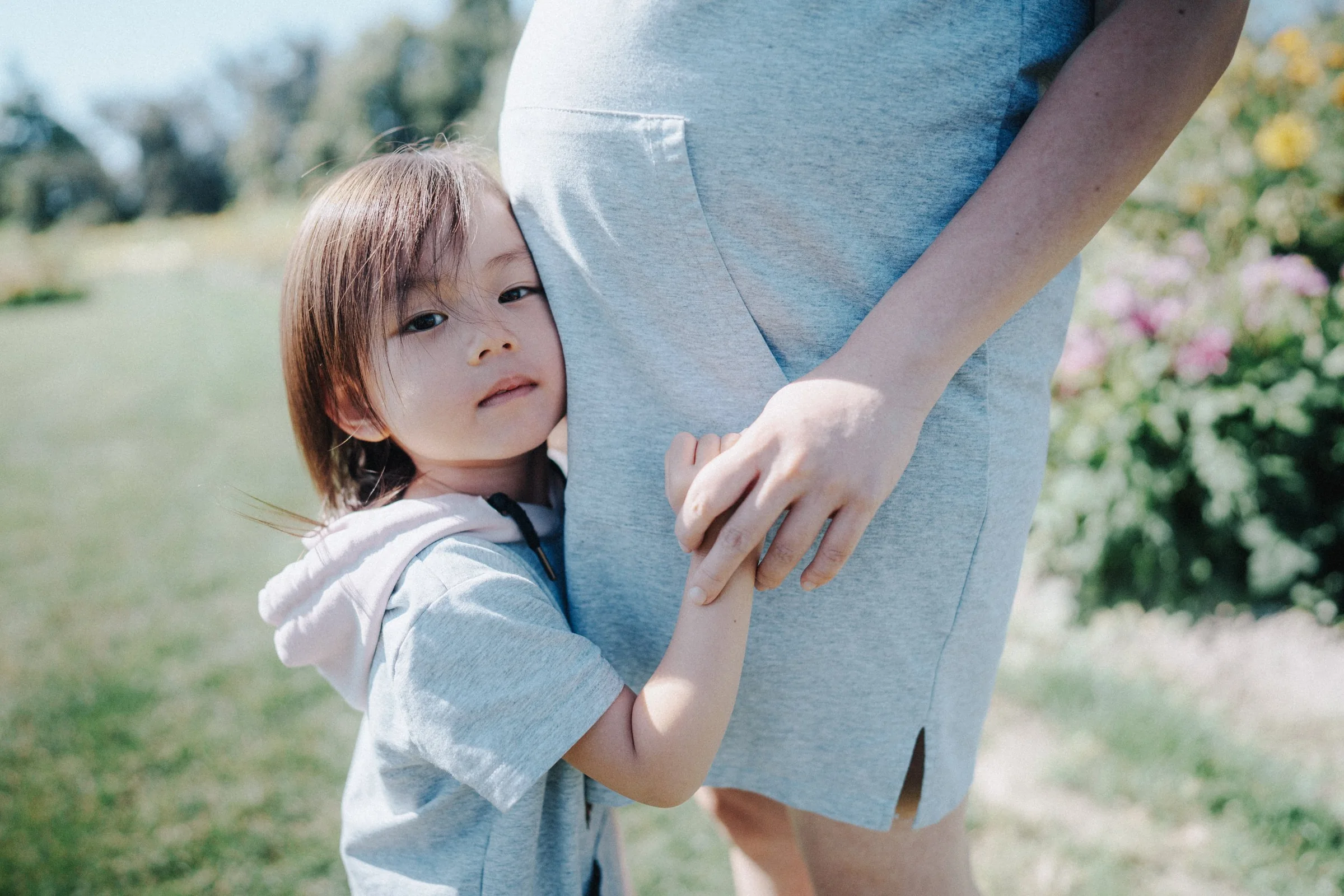 A young girl hugging her mother's leg in a sunny garden