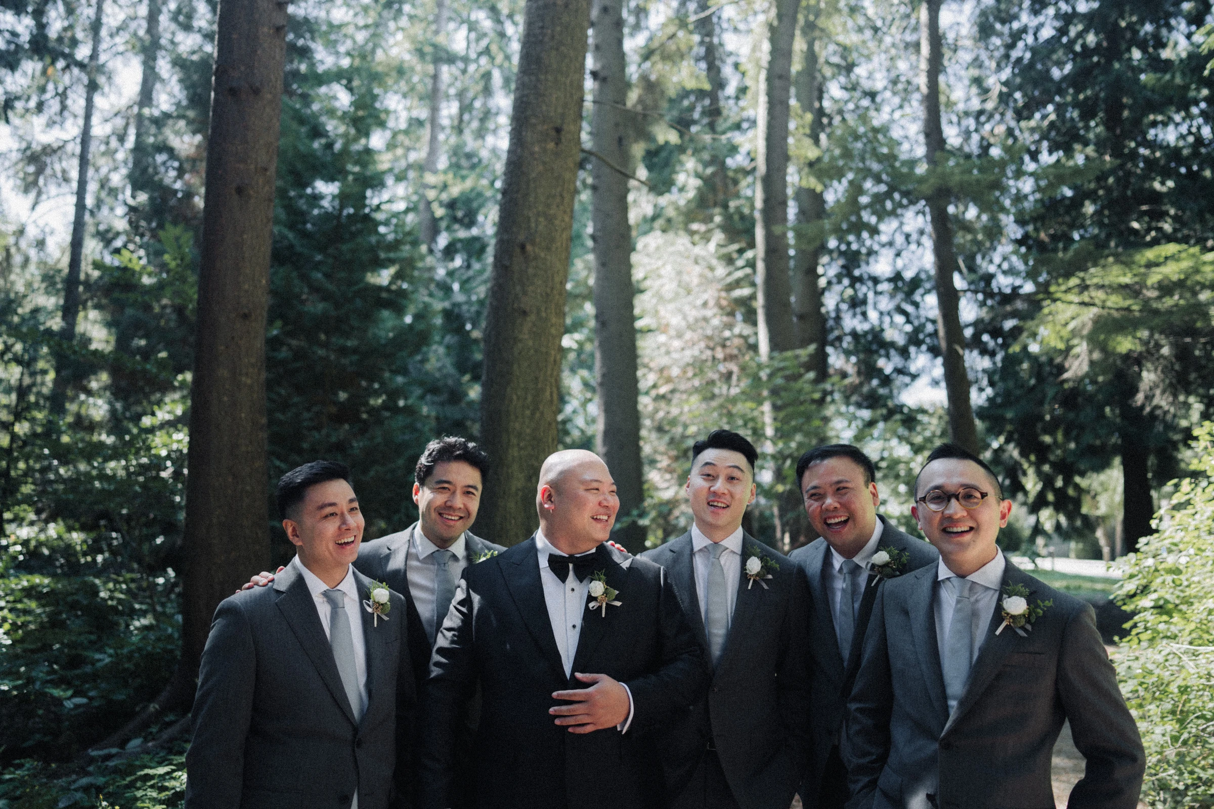 Groom and groomsmen standing shoulder to shoulder beneath the trees.