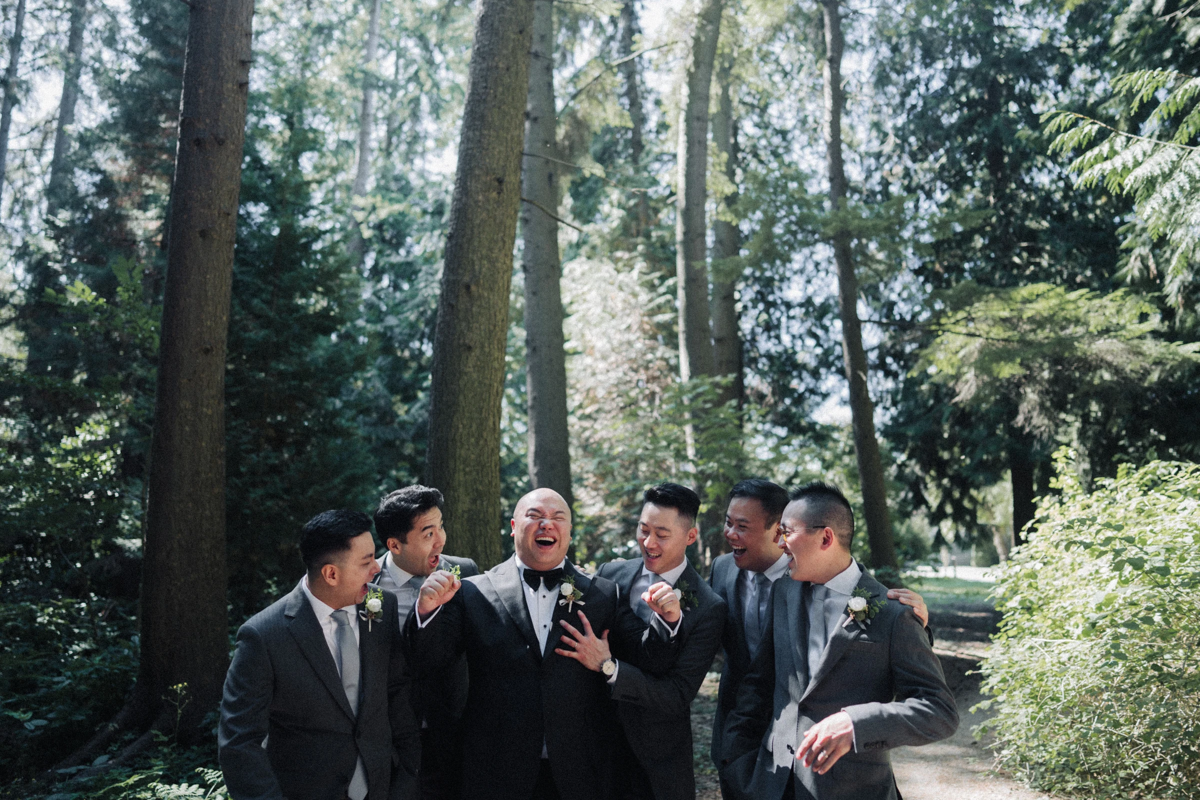 Groomsmen laughing together in a patch of woodland shade.