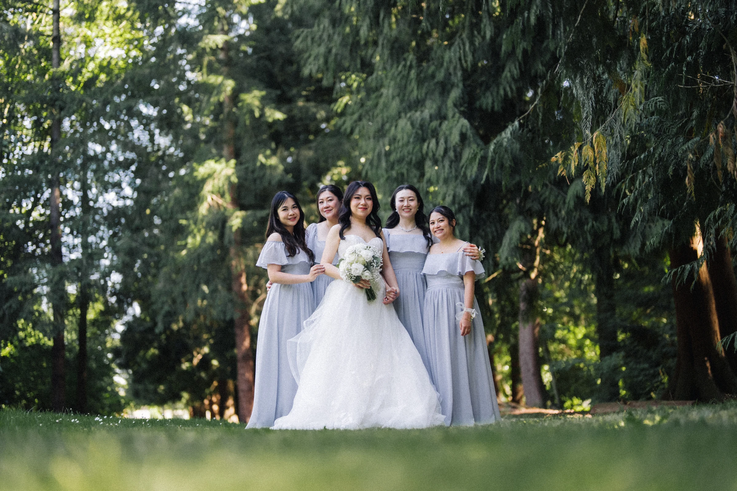 Bride standing with bridesmaids in pale blue dresses beneath the trees.