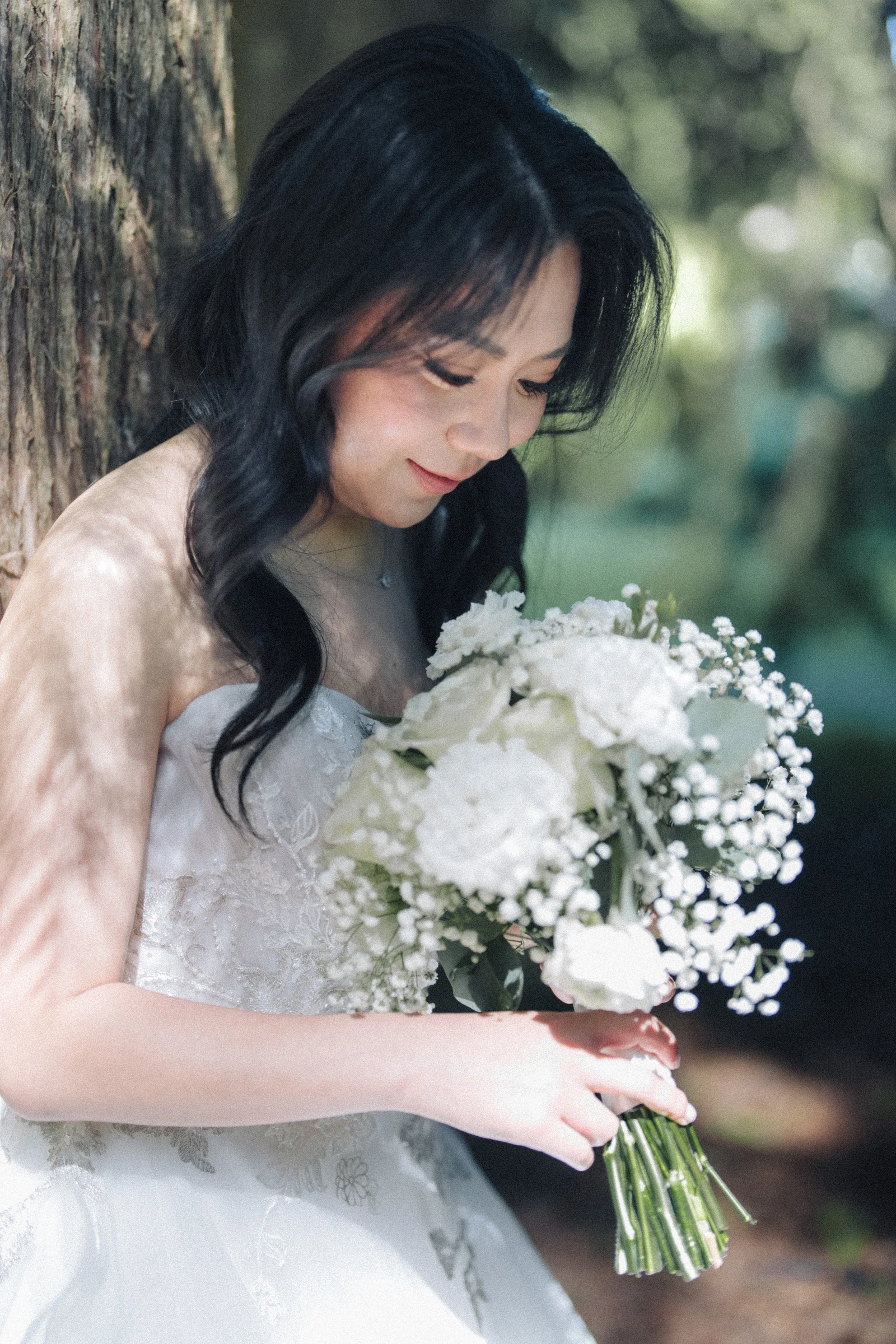 Bride looking down towards her white bouquet in the woodland shade.