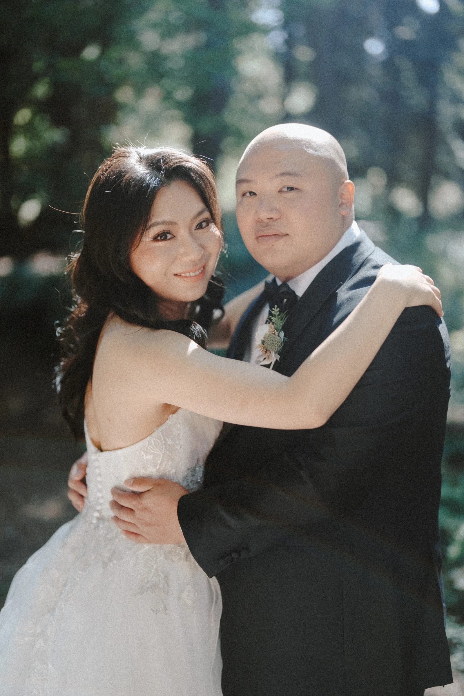 Couple embracing beneath the trees, with the bride looking towards the camera.