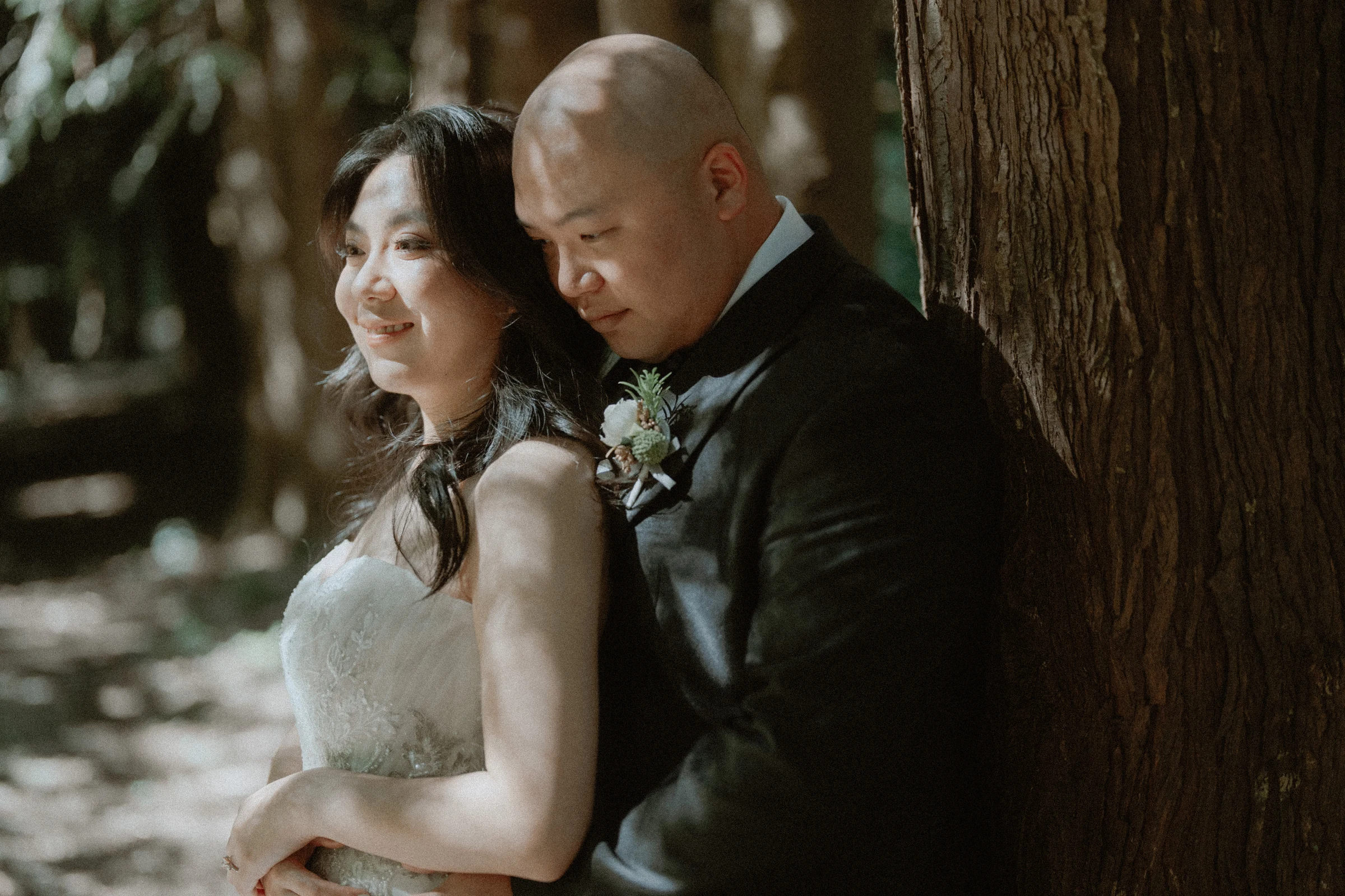 Couple leaning together against a broad tree trunk.