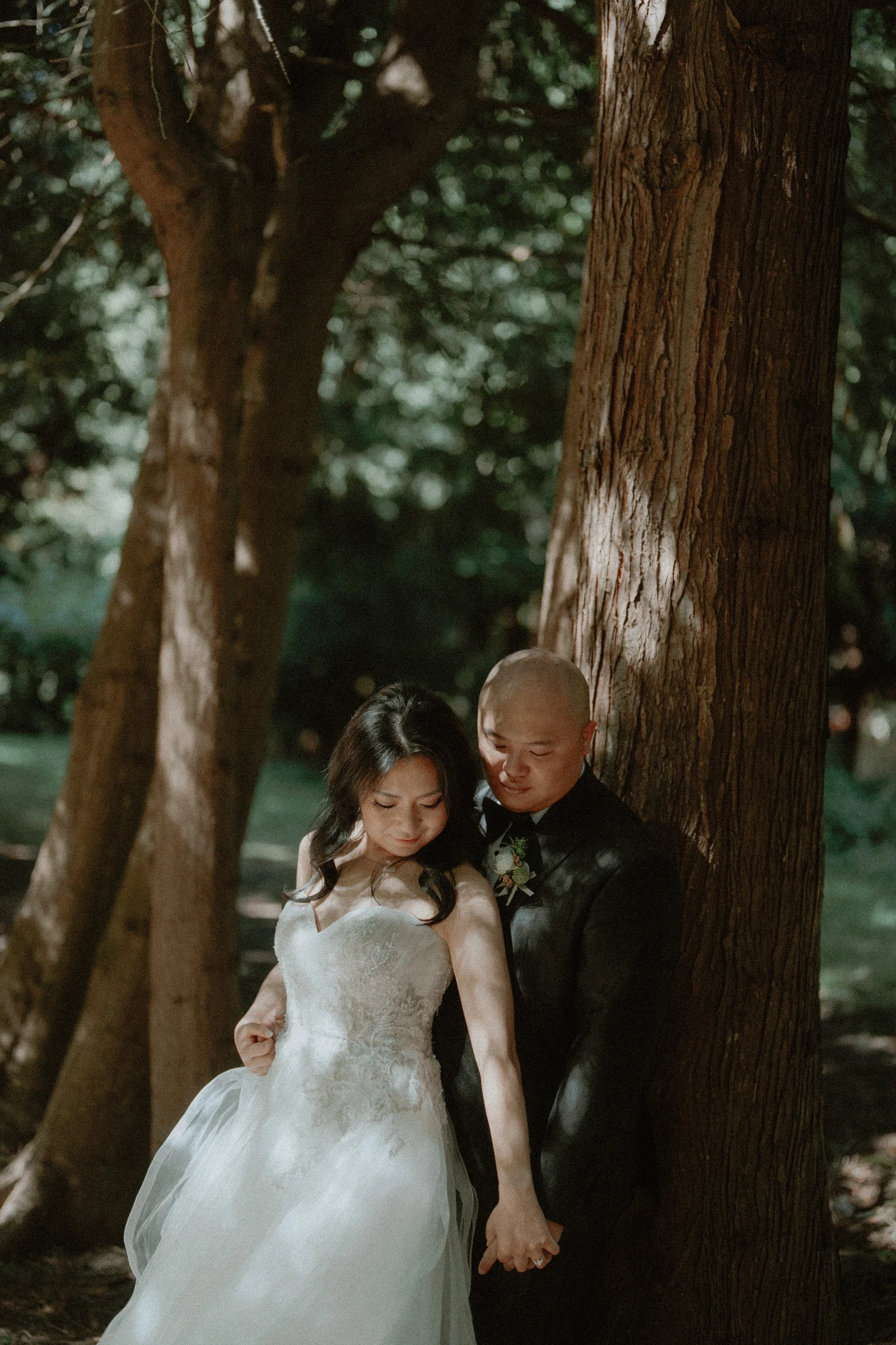 Bride and groom leaning together beneath a tall woodland trunk.