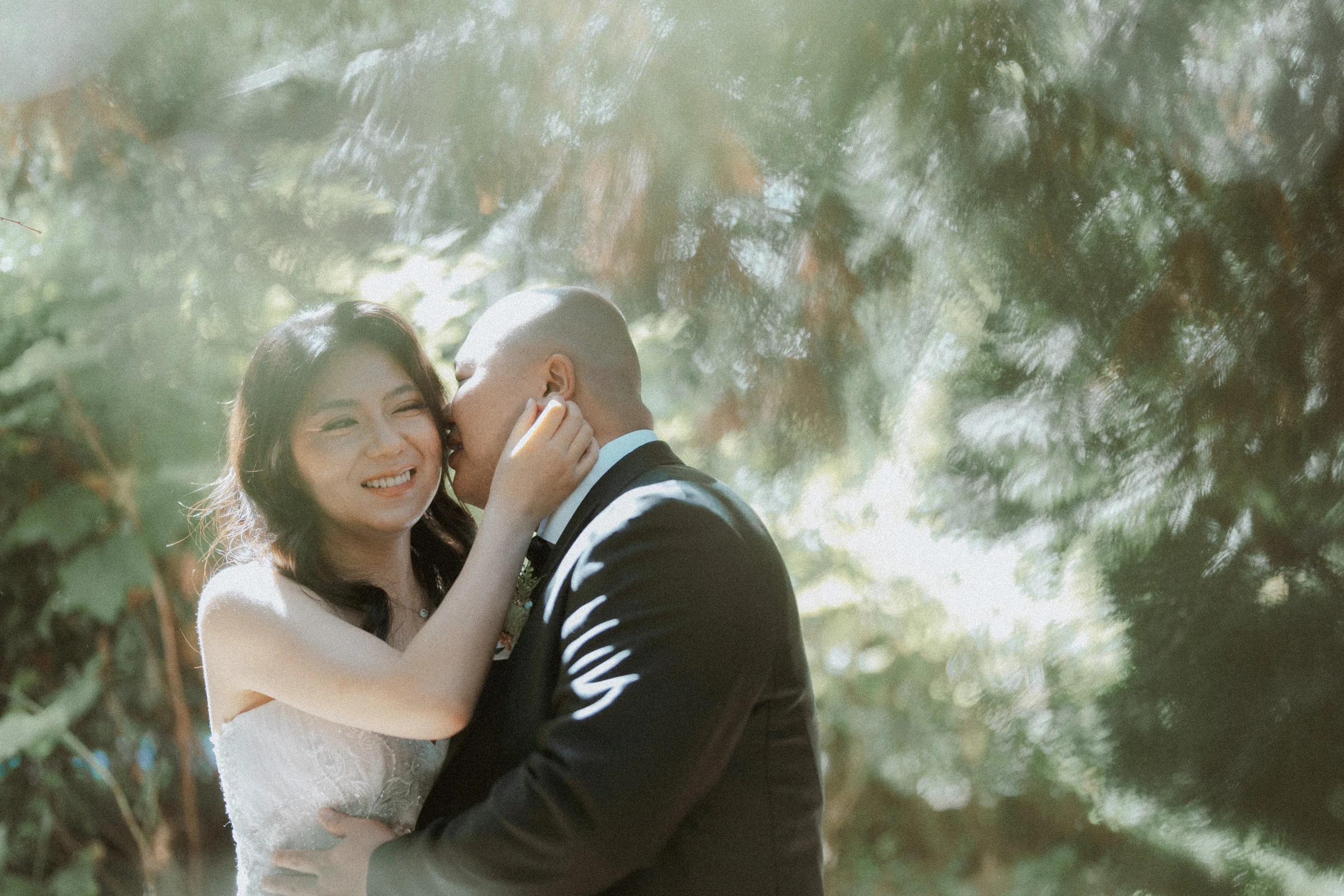 Bride smiling as the groom leans towards her in the sunlight.