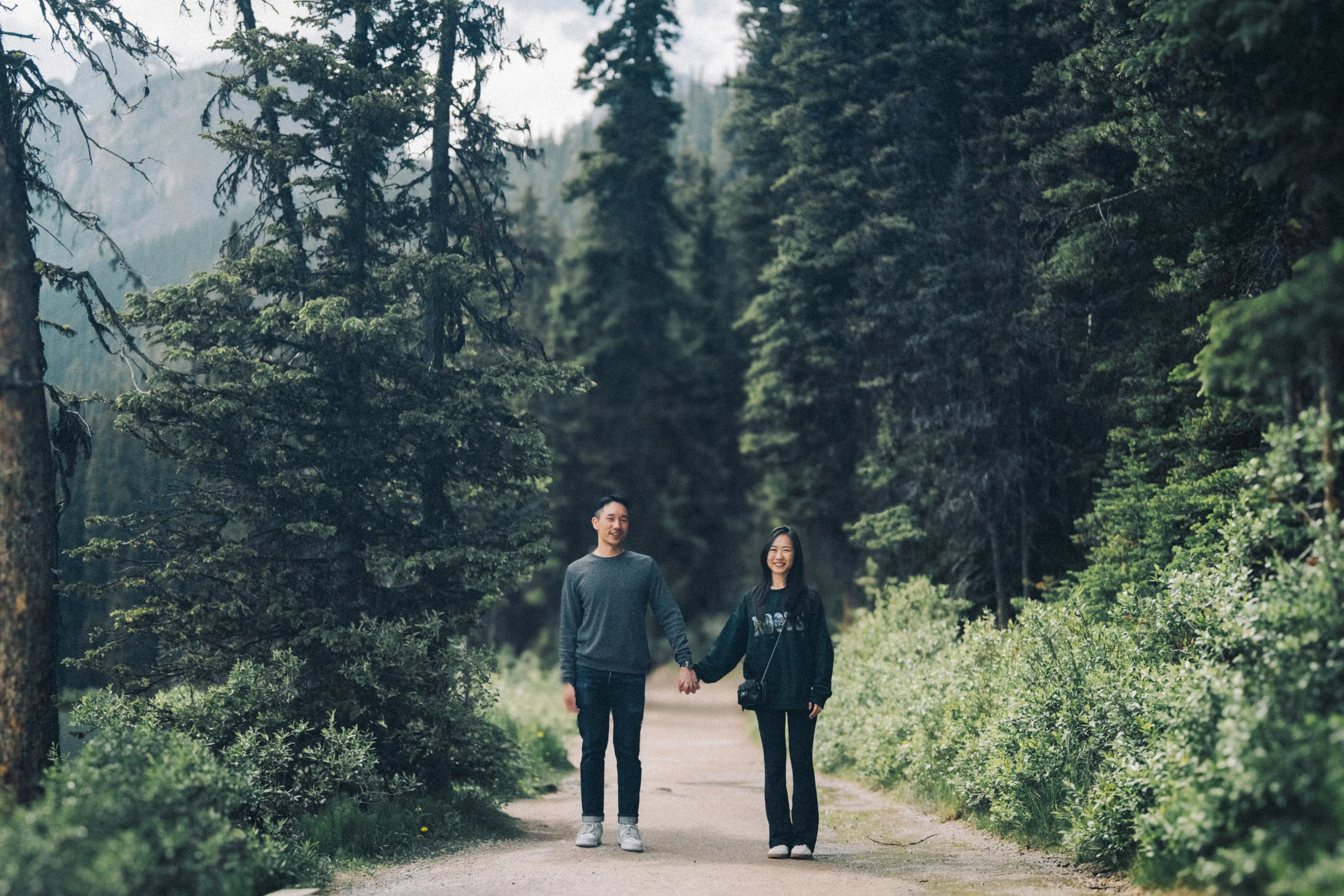 Rachael and Desmond hold hands on a path among evergreen trees at Lake Louise in Banff National Park.