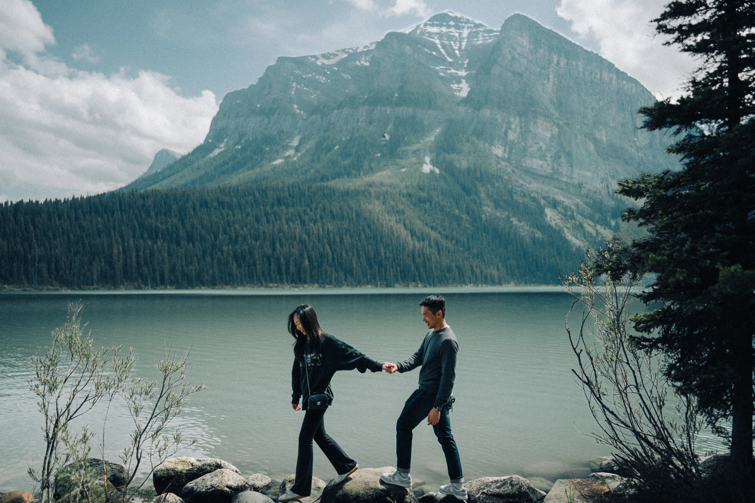 Rachael and Desmond walk hand in hand across rocks beside Lake Louise in Banff National Park, with mountains behind them.