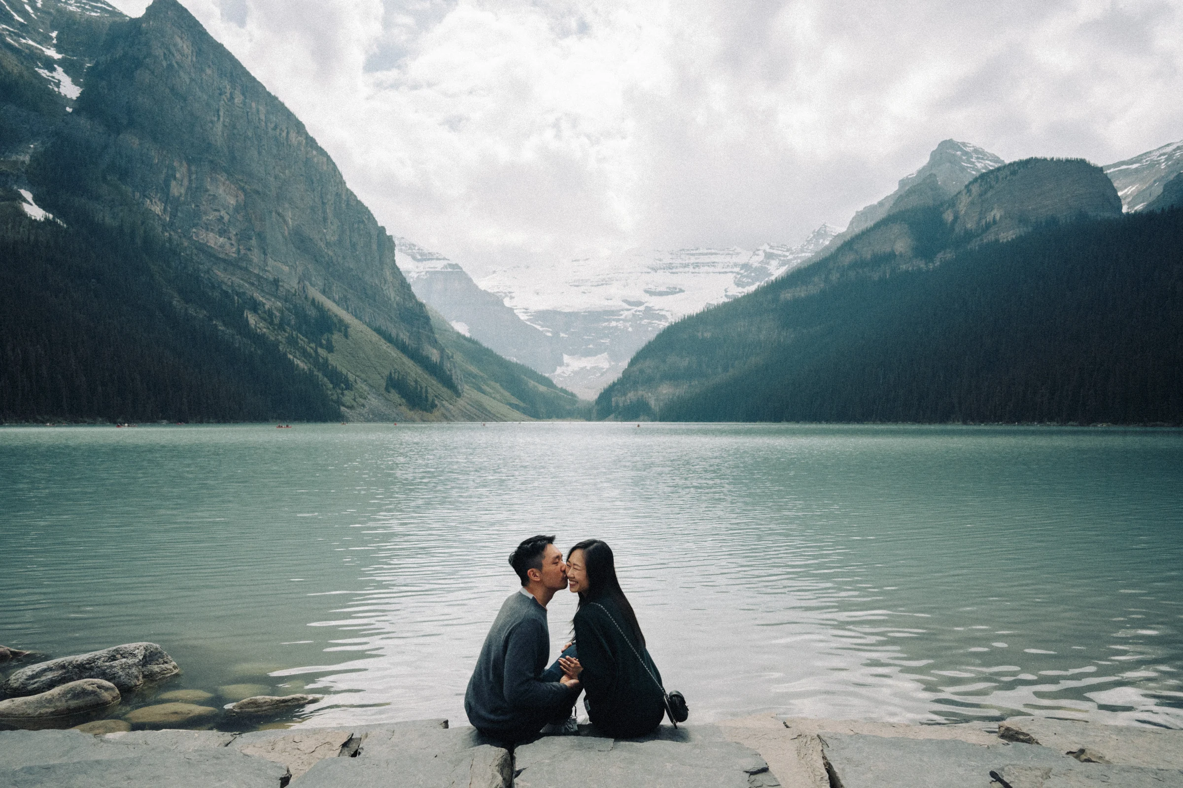 Rachael and Desmond sit and kiss on rocks at Lake Louise in Banff National Park beneath the mountains.