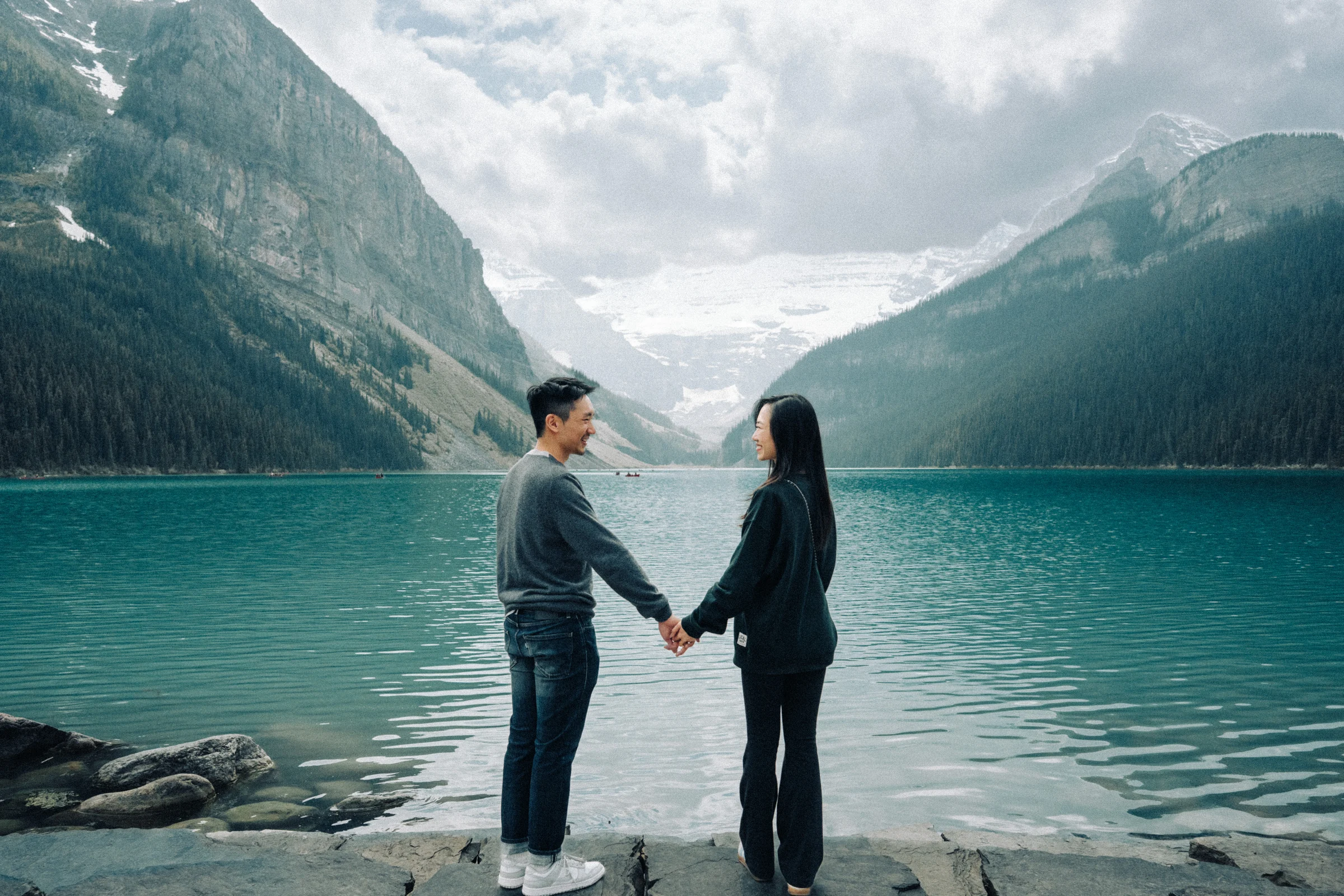 Rachael and Desmond stand hand in hand on the rocky shore of Lake Louise in Banff National Park.