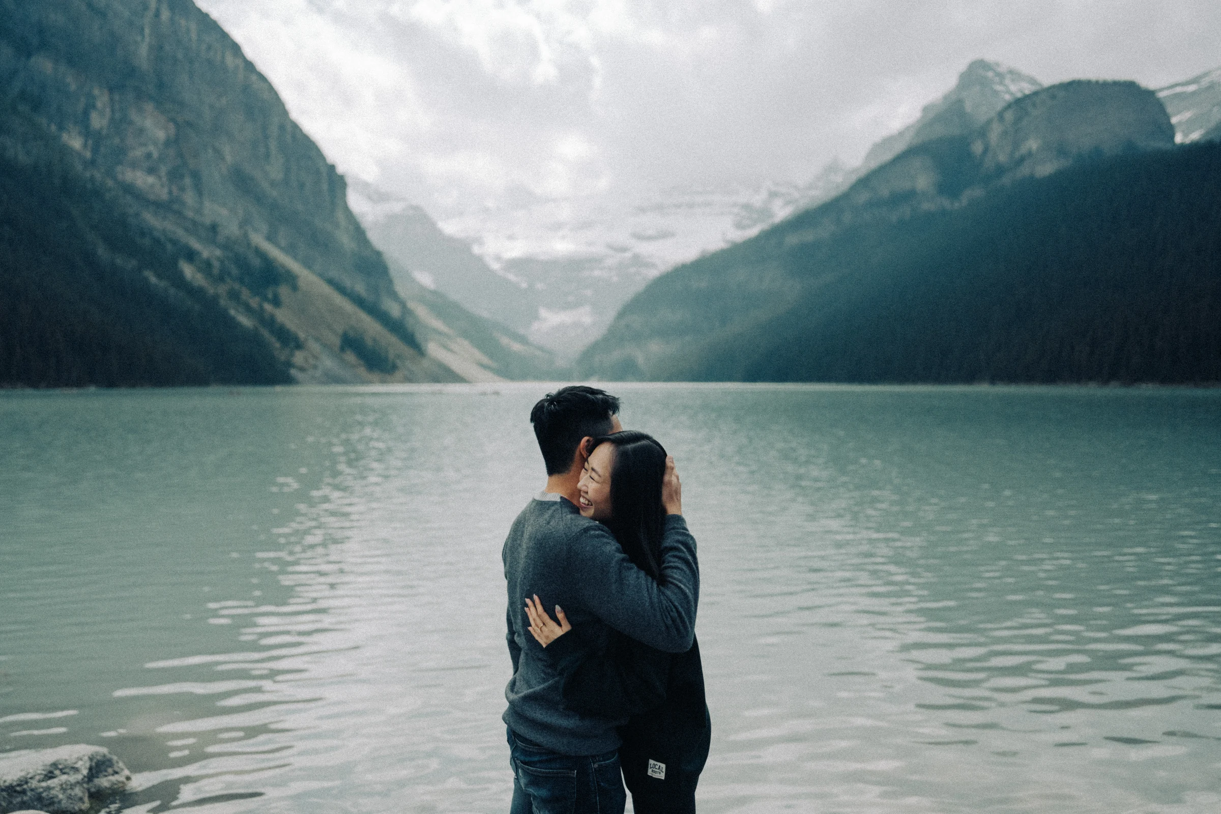 Rachael and Desmond embrace on the shore of Lake Louise in Banff National Park beneath the mountain backdrop.
