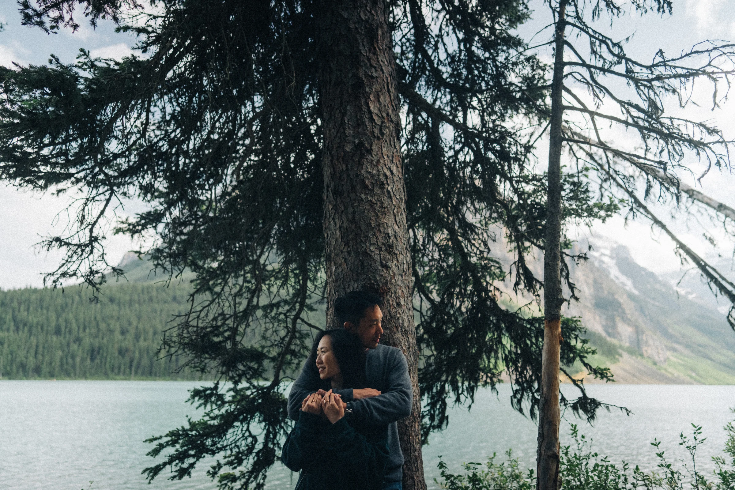 Desmond embraces Rachael beneath evergreen branches beside Lake Louise in Banff National Park.