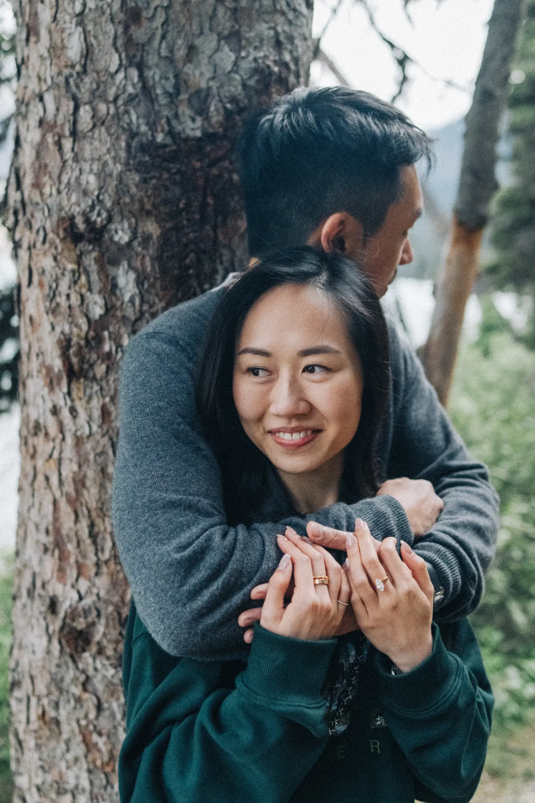 Rachael looks toward the camera as Desmond embraces her beside a tree, at Lake Louise in Banff National Park.