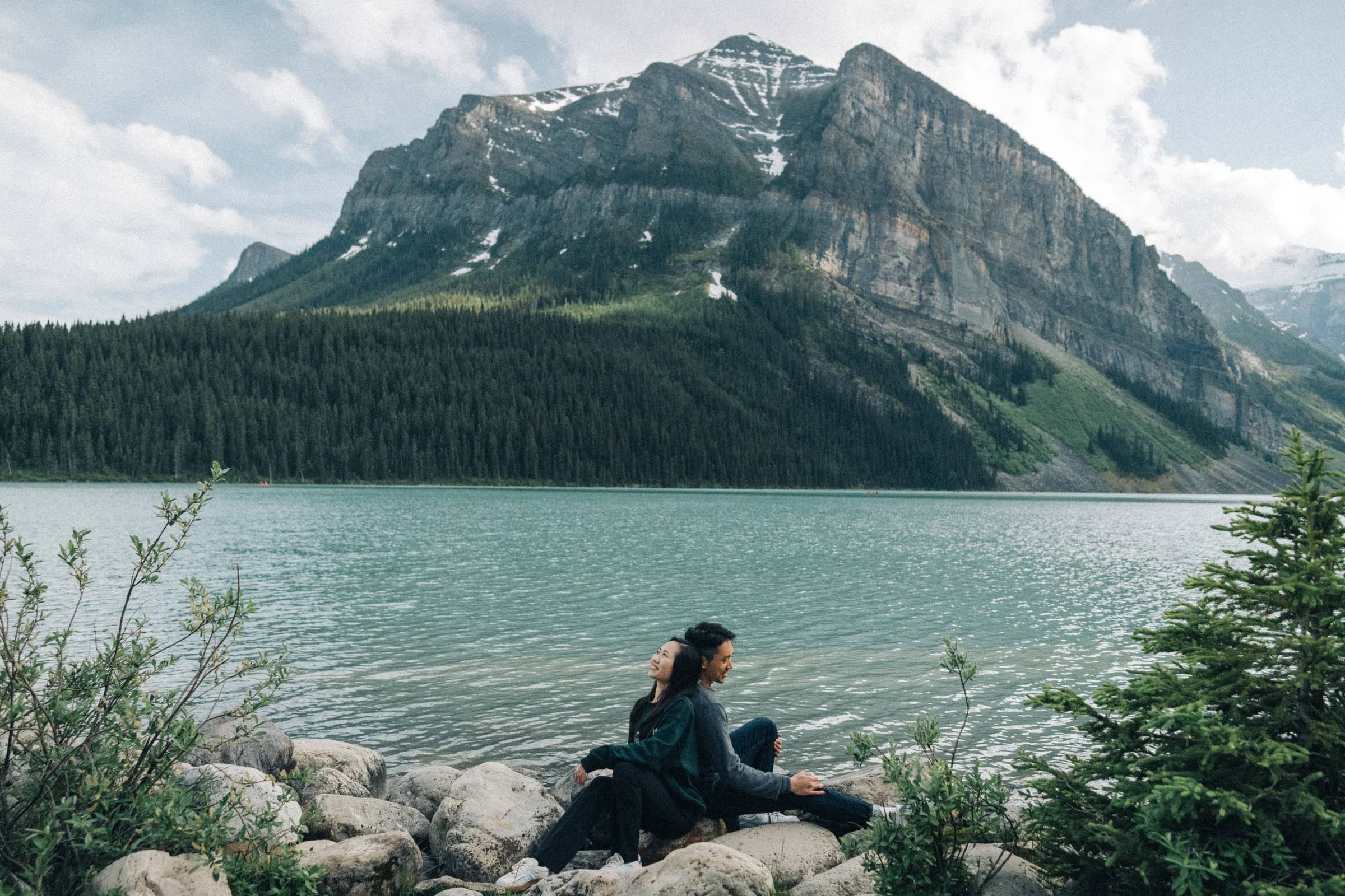 Rachael and Desmond sit together on lakeside rocks beneath a mountain view, at Lake Louise in Banff National Park.