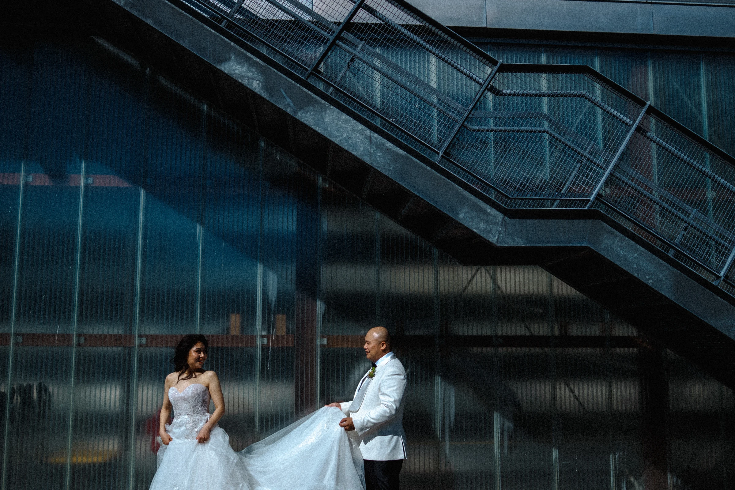 Bride and groom standing apart beneath the diagonal steel stairs.