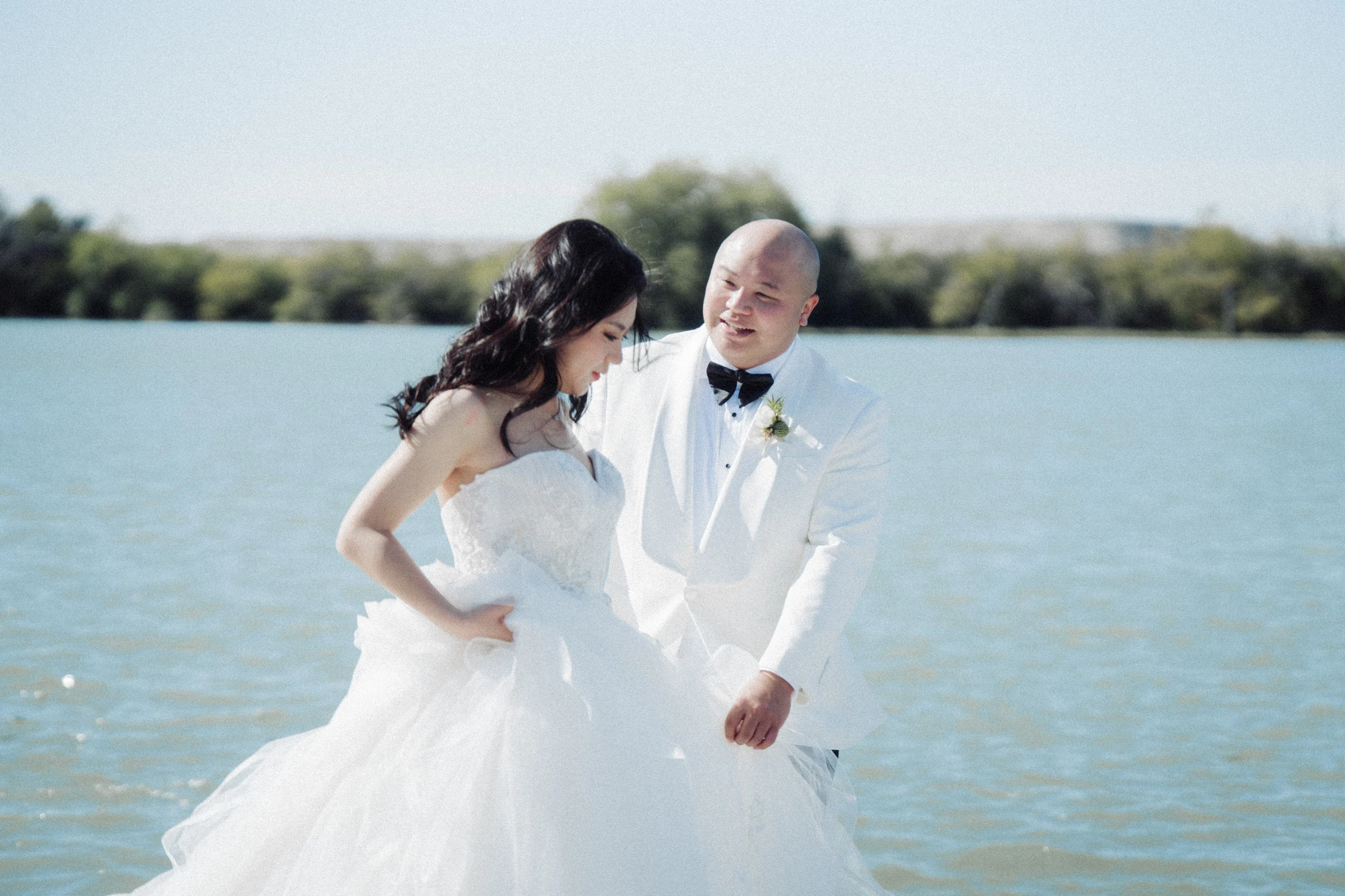Bride and groom laughing beside the river, with the dress gathered in motion.