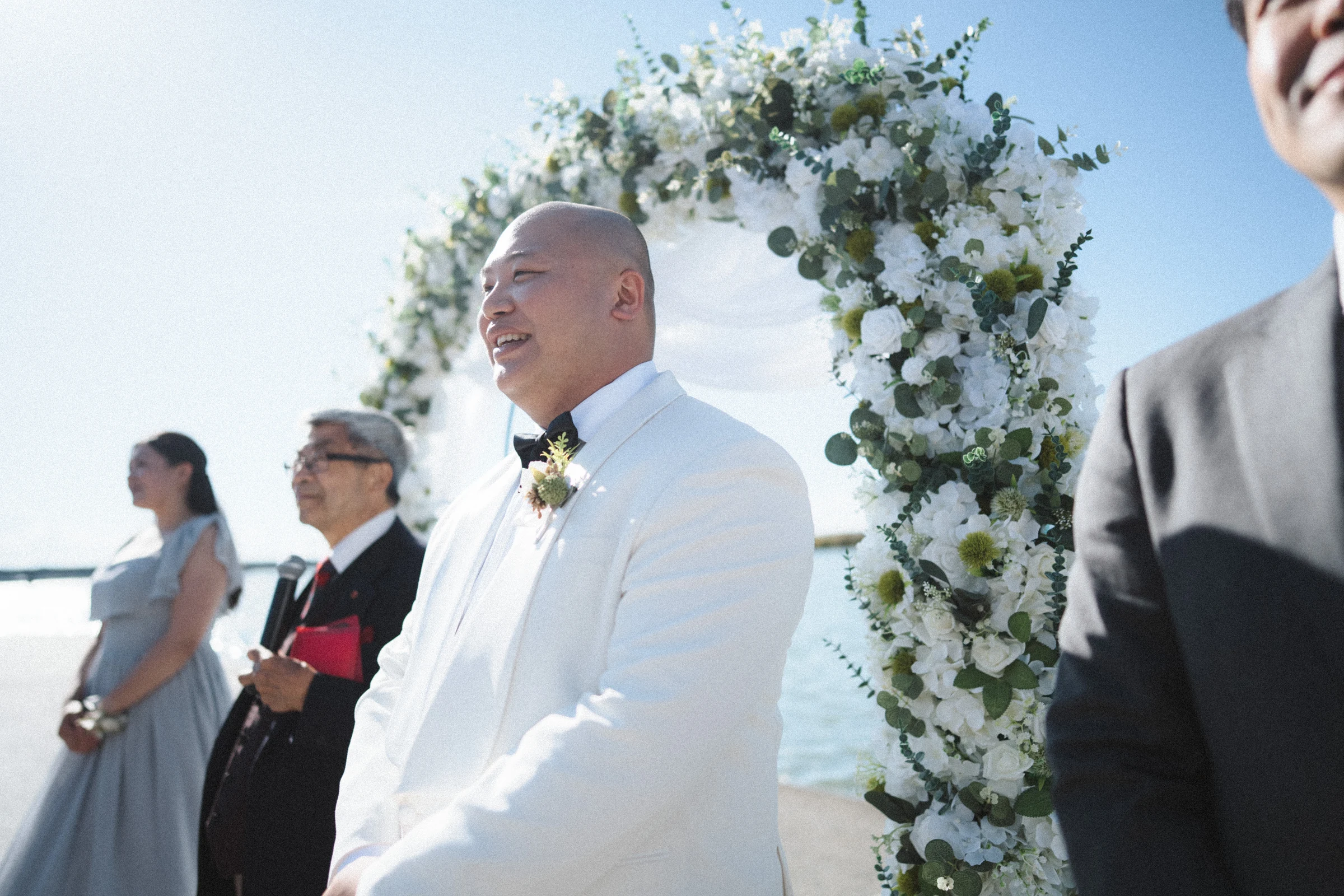 Groom waiting beside the flower arch in a white jacket.