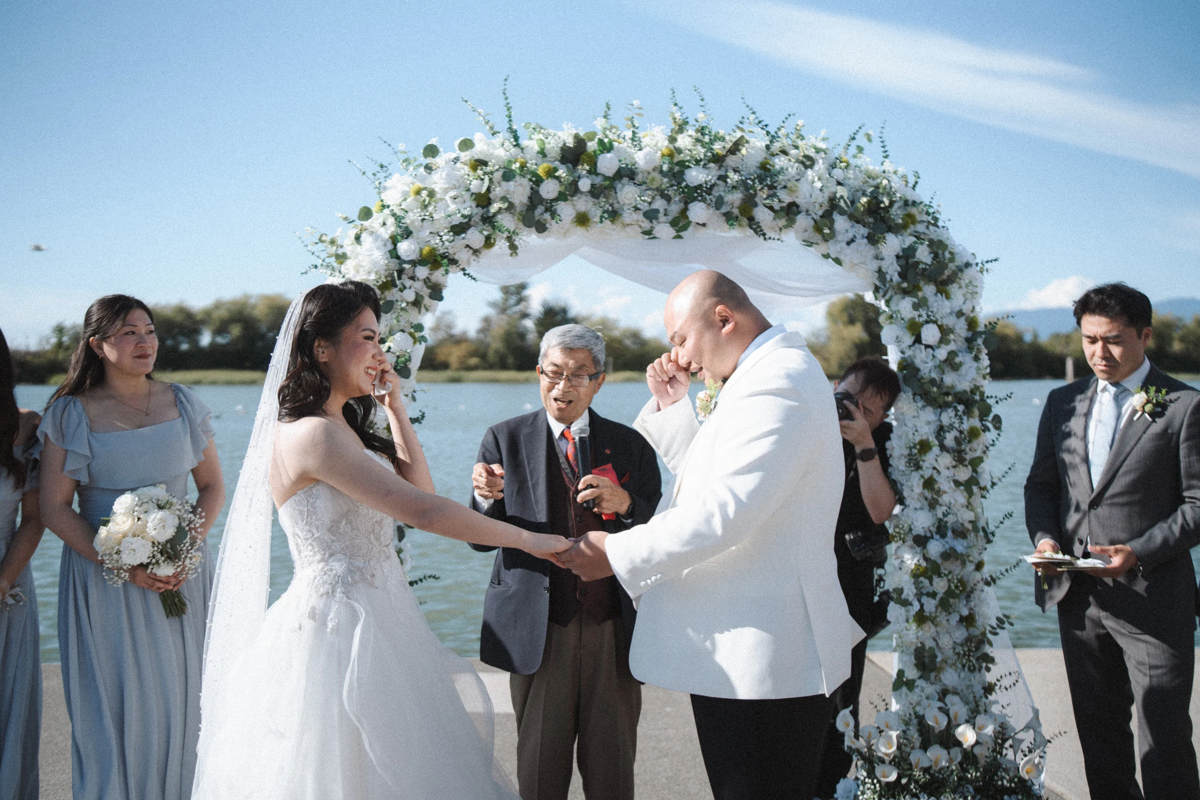 Couple holding hands beneath the white flower arch beside the Fraser River.