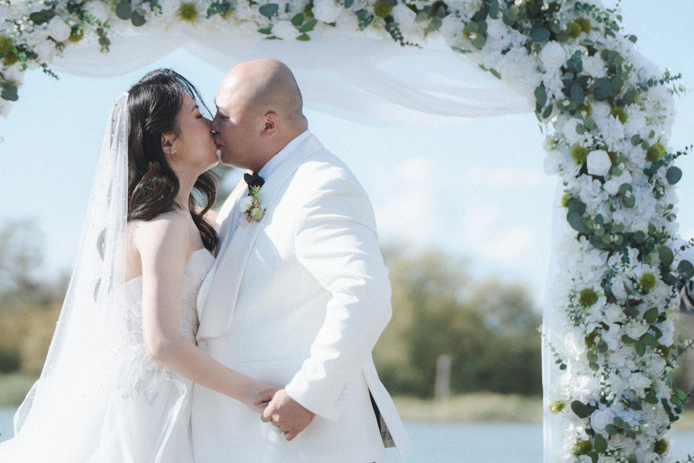Bride and groom kissing beneath the white flower arch.