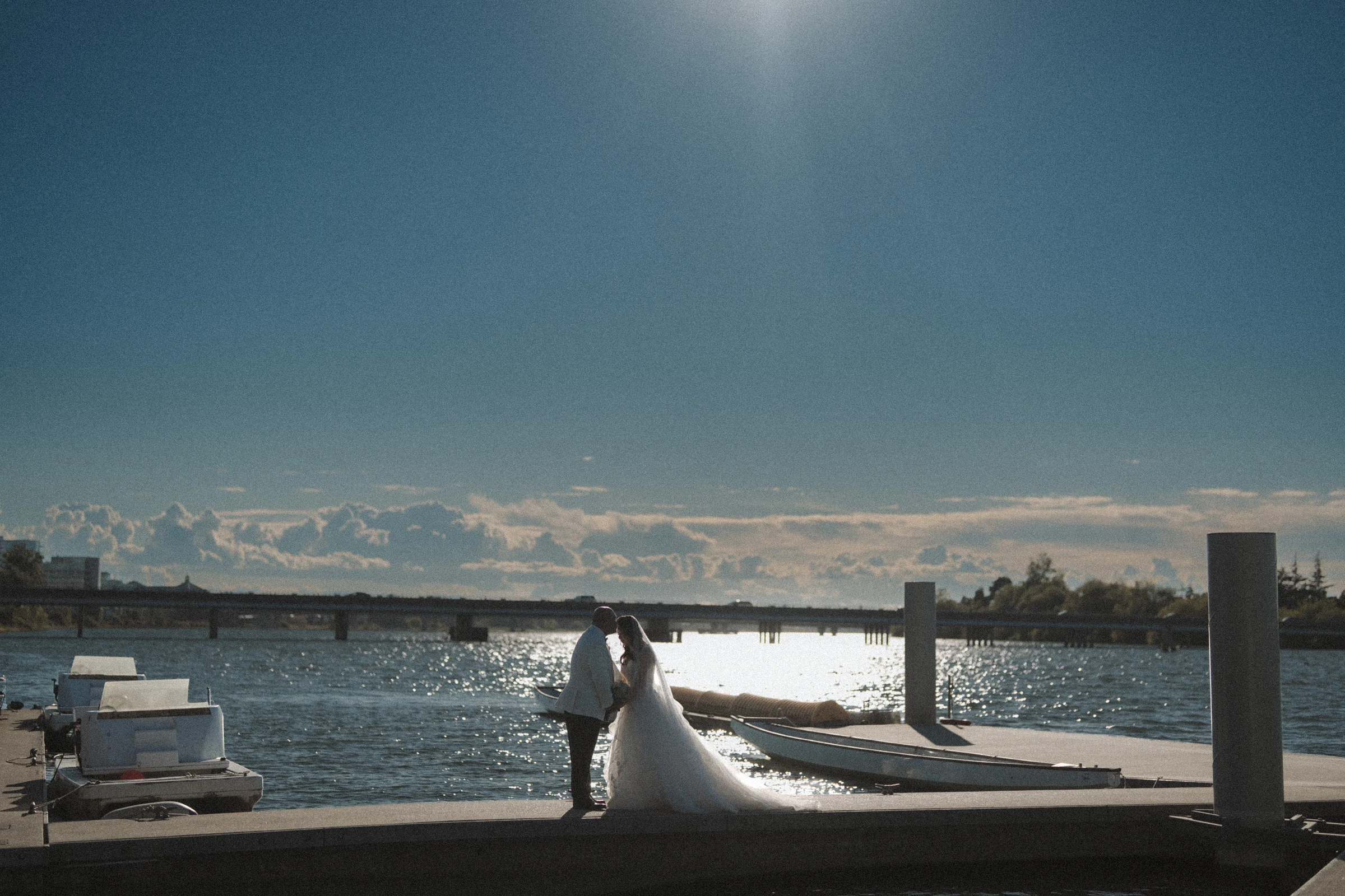 Wide dockside portrait of the couple, with small boats and bright water behind them.