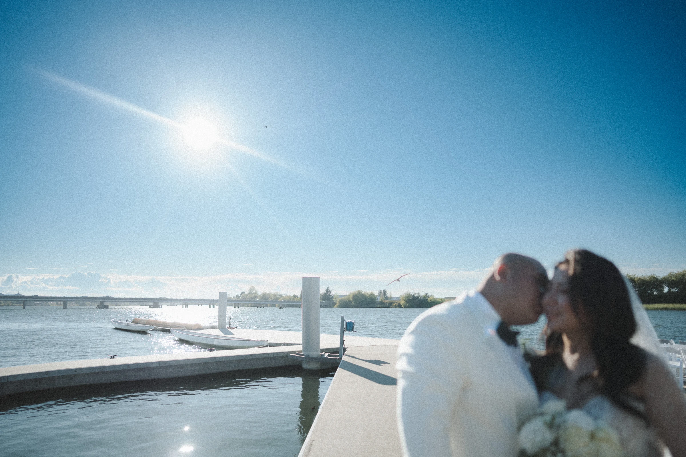 Bride and groom kissing beside the river in strong afternoon sunlight.