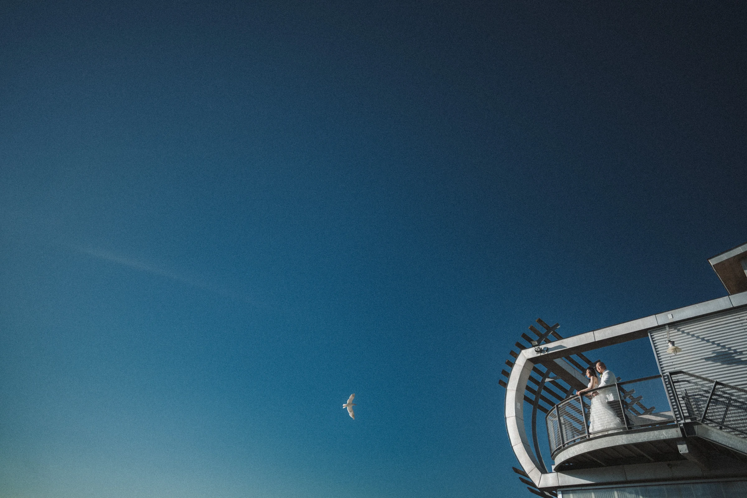 Bride on the upper boathouse balcony beneath a wide blue sky.