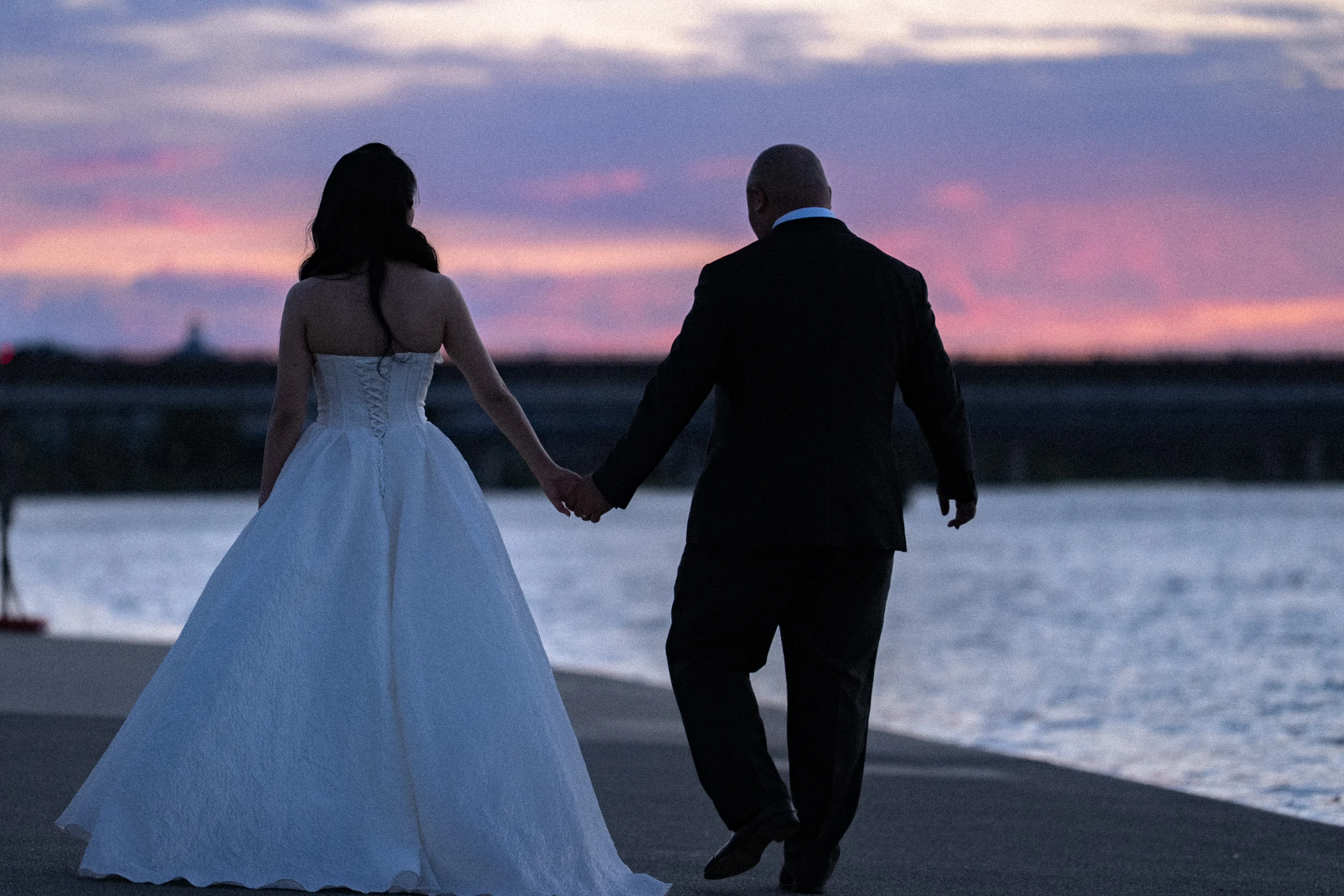 Bride and groom walking hand in hand towards the river at dusk.