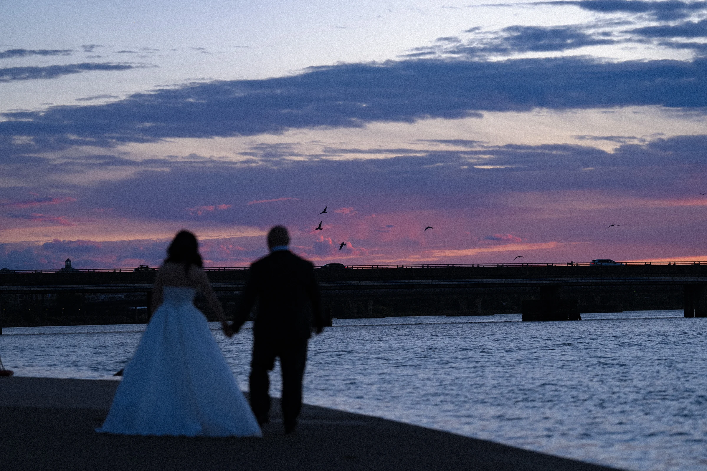 Couple standing beside the river under layered pink and blue clouds.