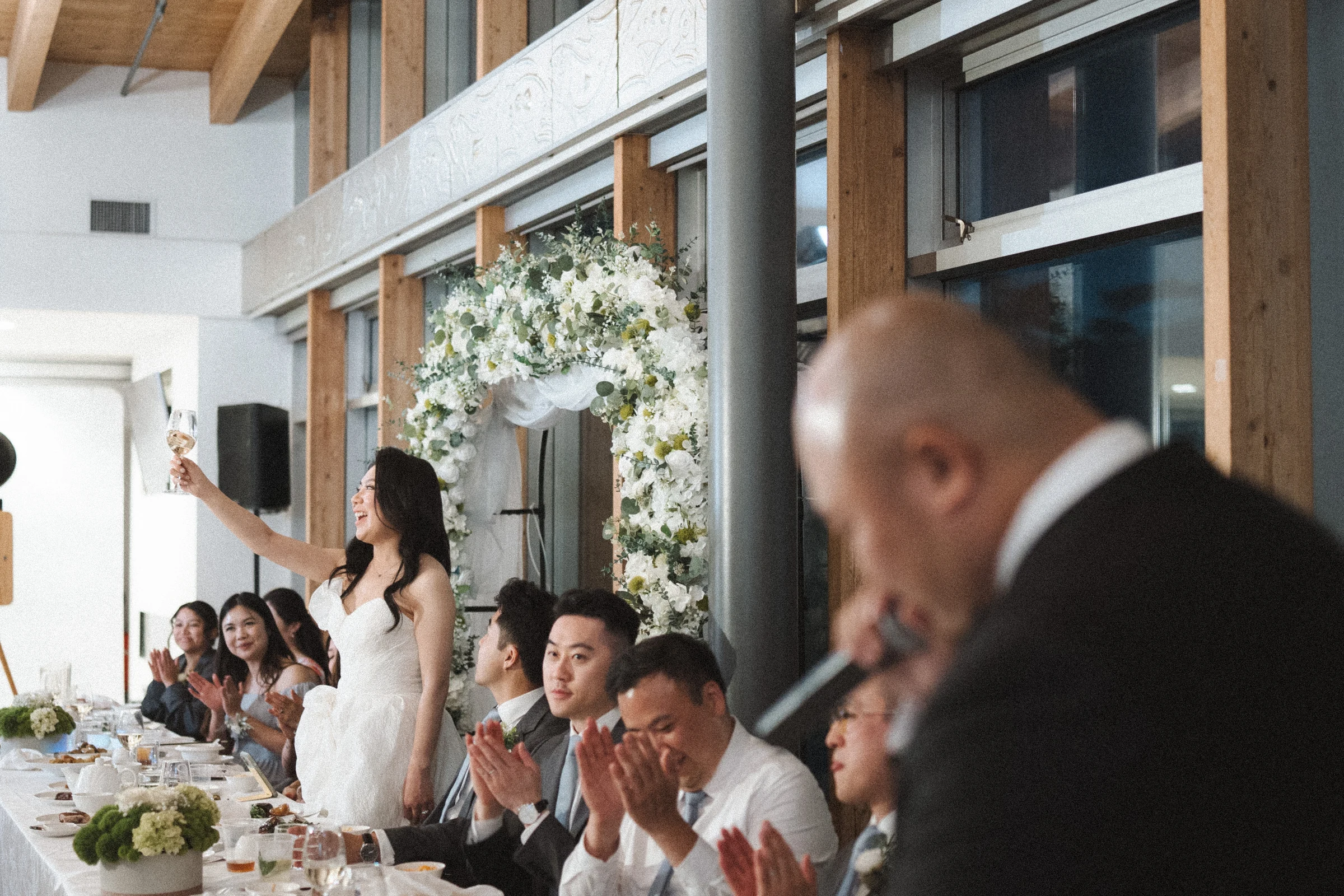 Couple seated by the flower arch as guests listen to a speech.