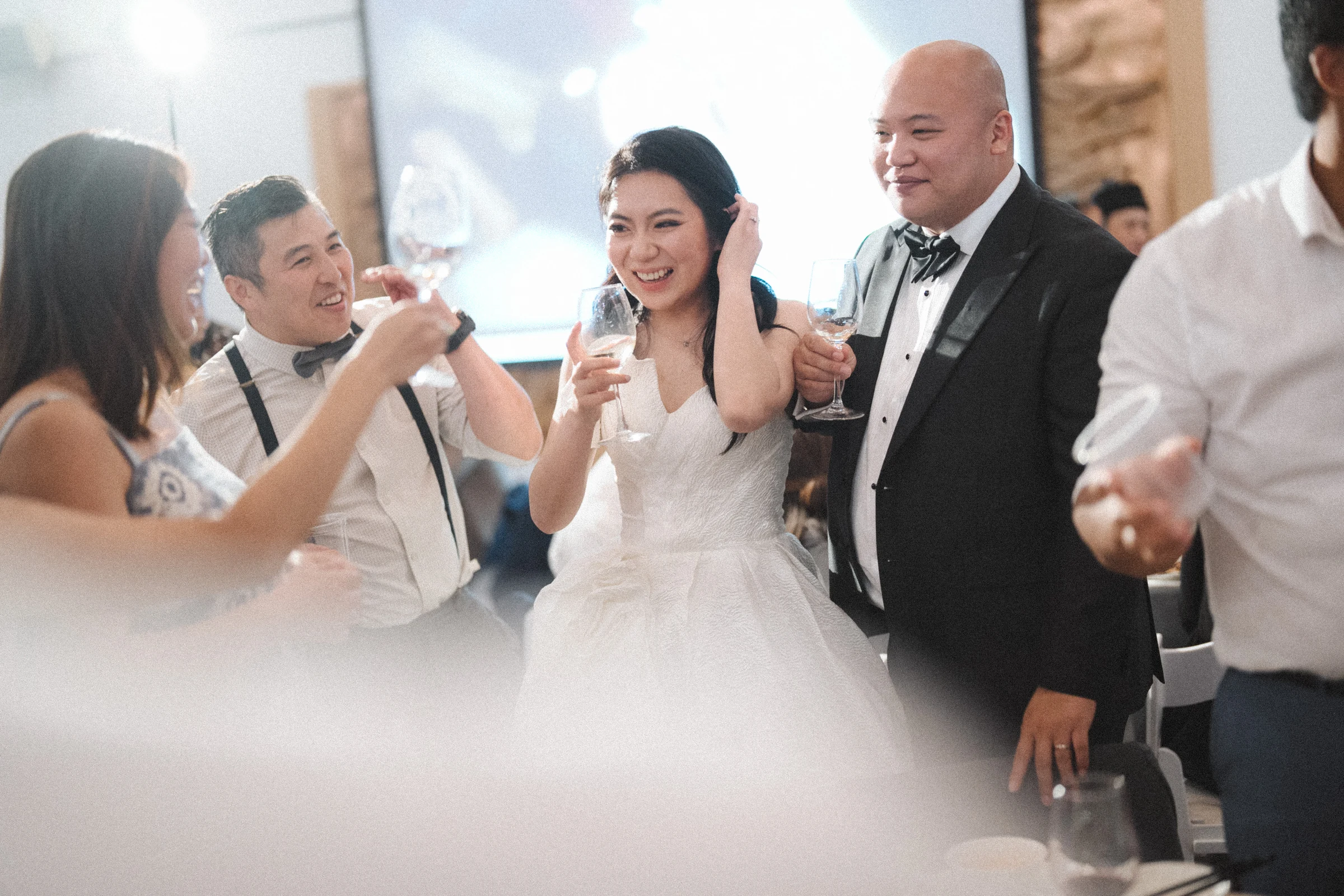 Bride and groom raising glasses while guests lean into the photograph.