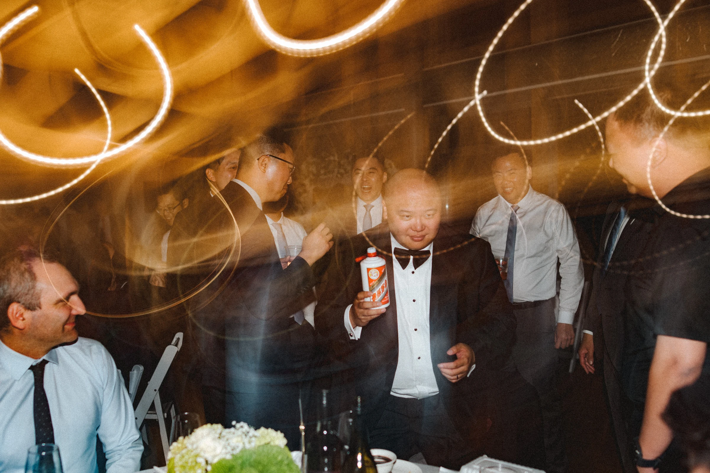 Groom dancing among friends beneath streaks of warm light.