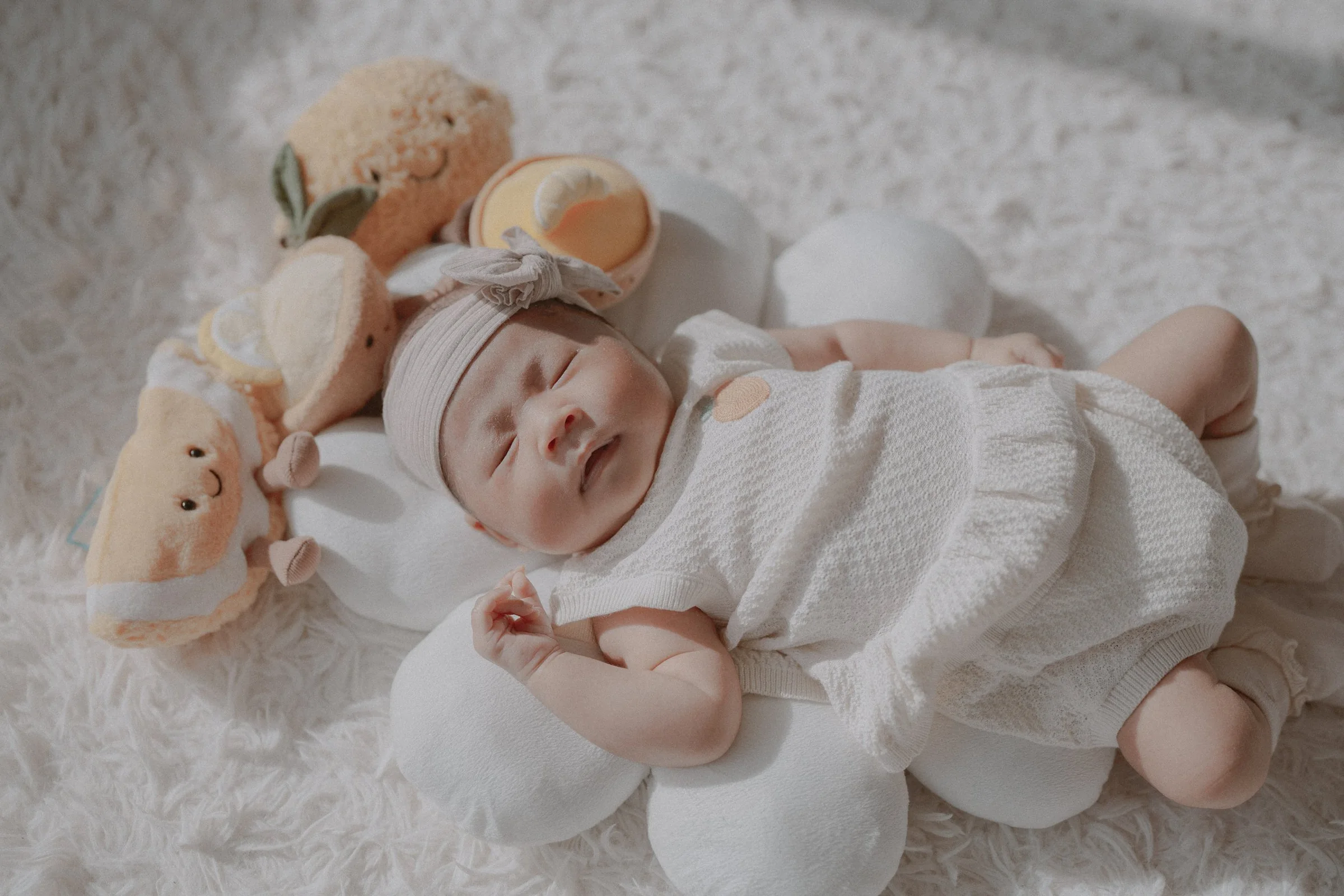 Newborn wearing a bow headband on a soft blanket beside cuddly toys.