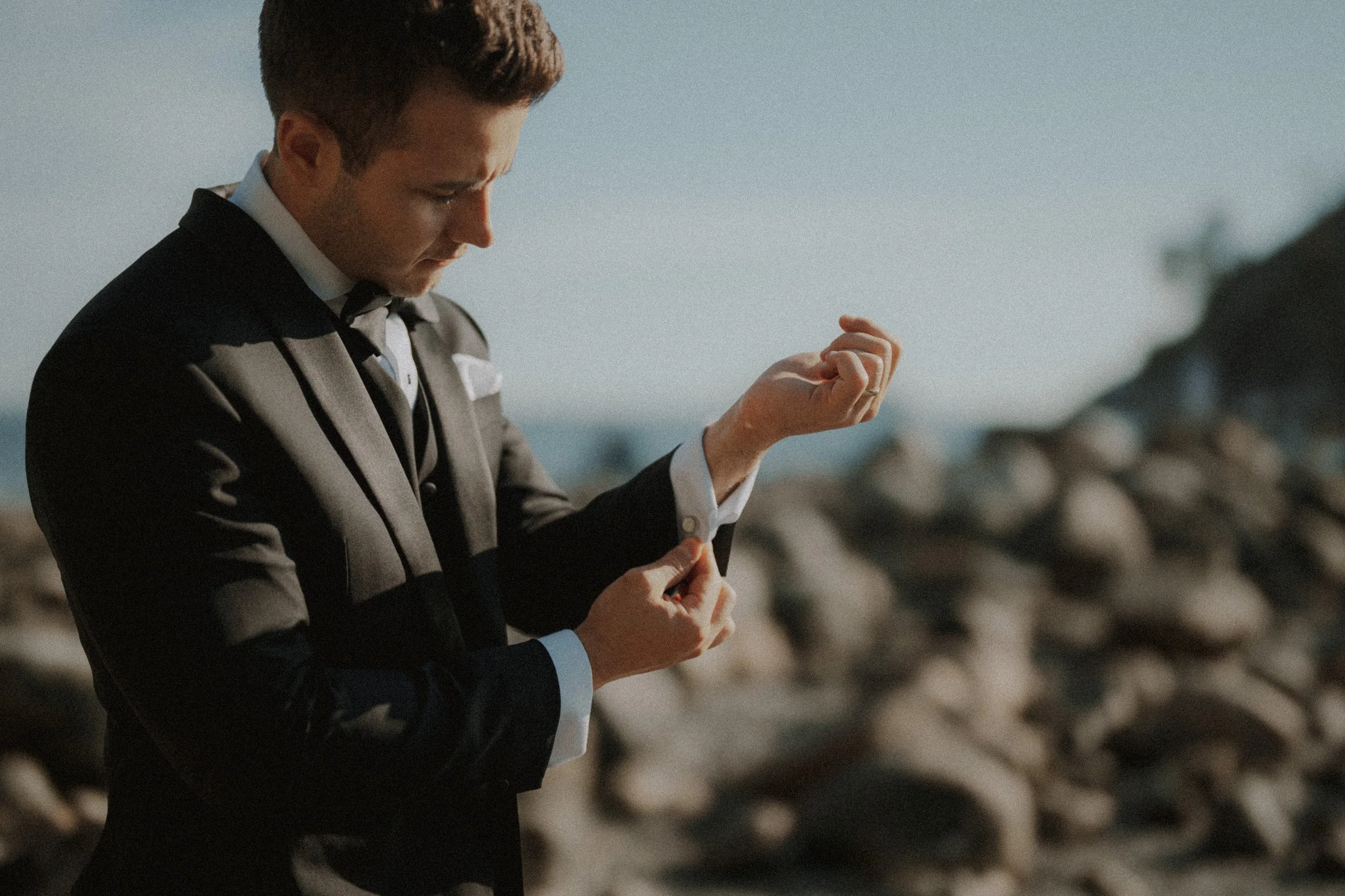 Groom adjusting his sleeve and cuff before a shoreline portrait.