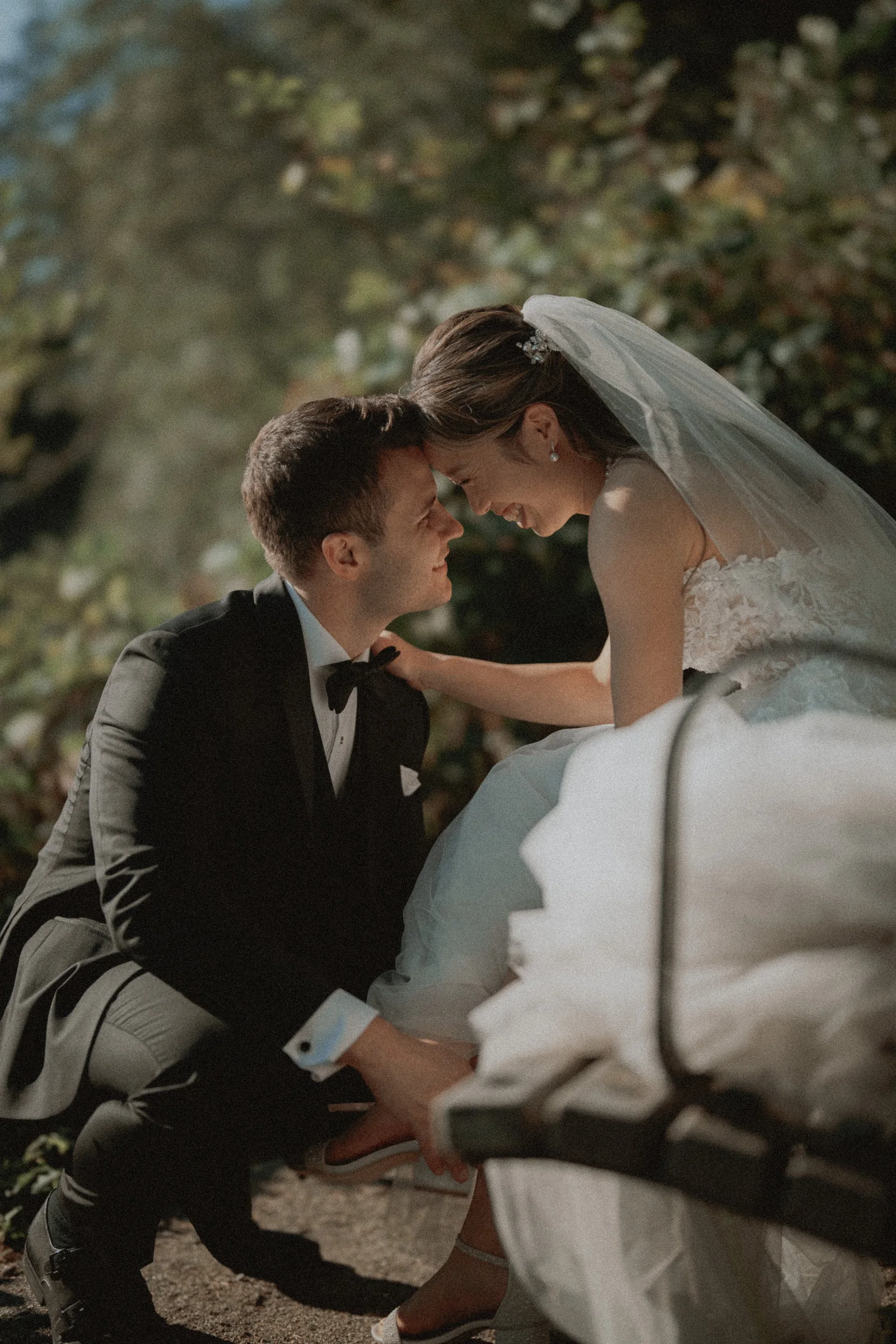 Groom crouching beside the bride as she sits on a shoreline bench.