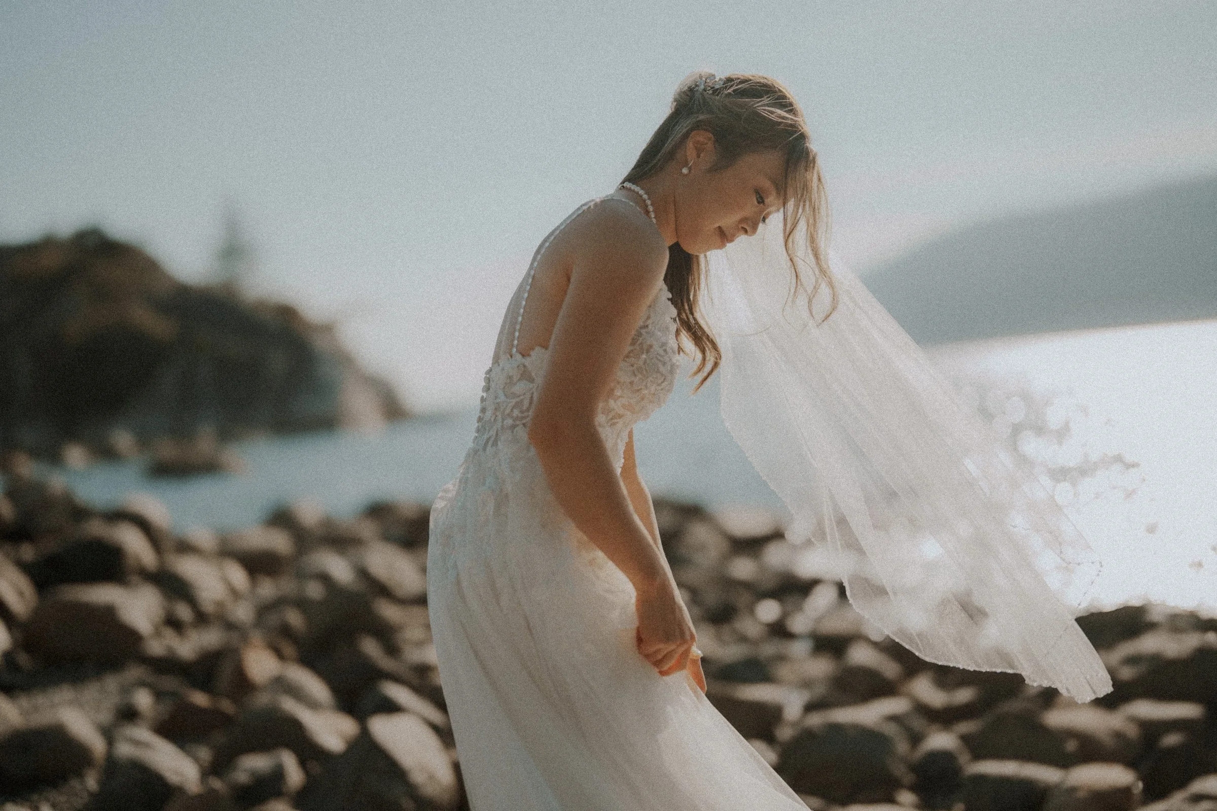 Bride's veil blowing in the sea breeze as she looks down at her gown.