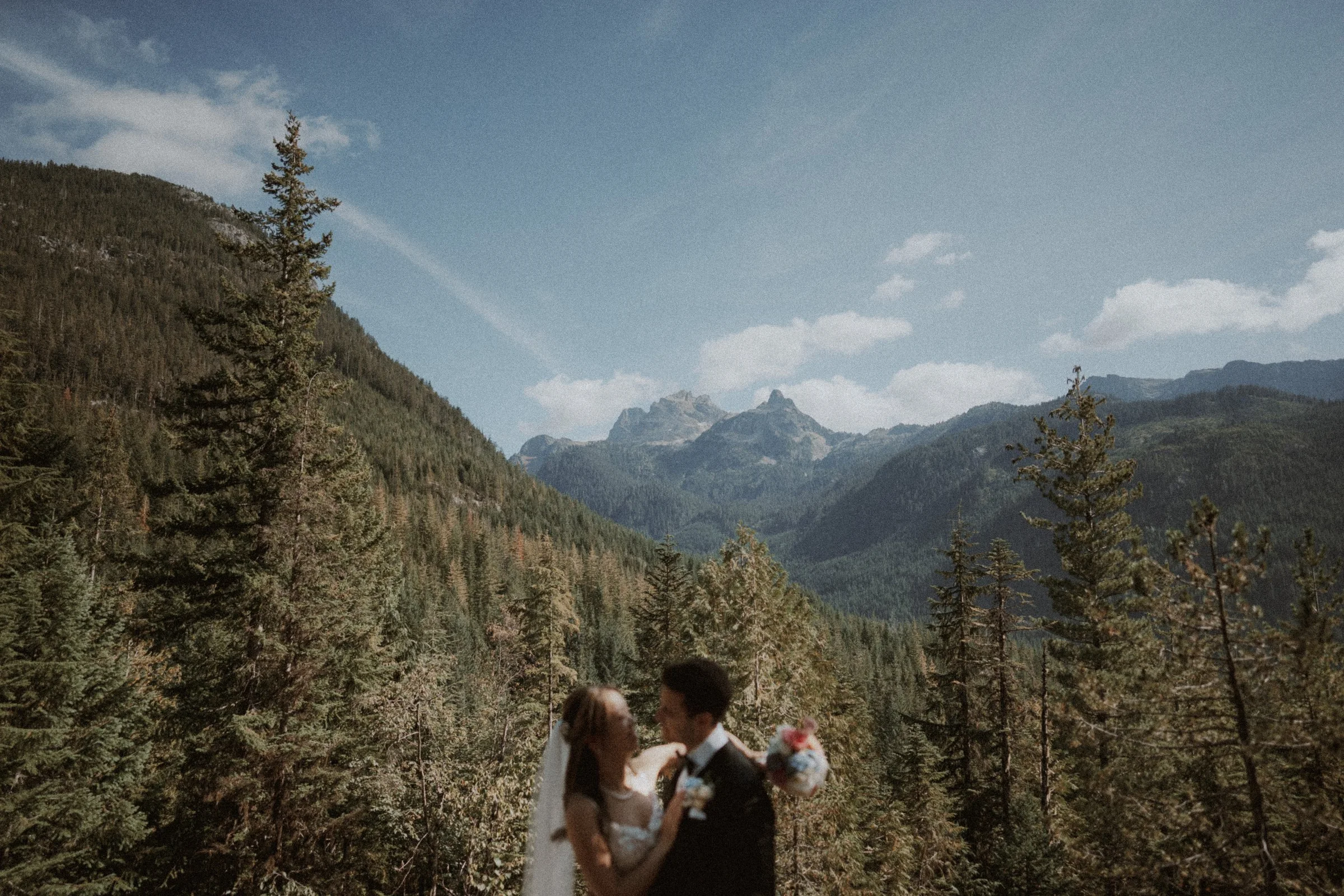 Couple embracing with a forested mountain valley stretching behind them.
