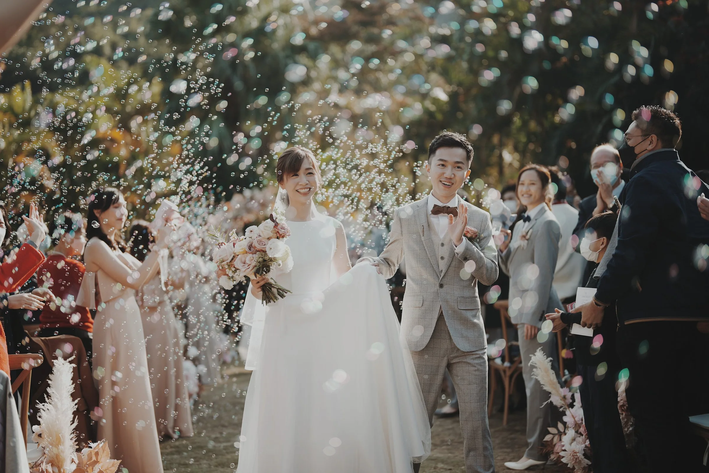 A newlywed couple walks hand in hand down the aisle outdoors, daughter holding bouquet, guests showering confetti, autumn trees in the background.