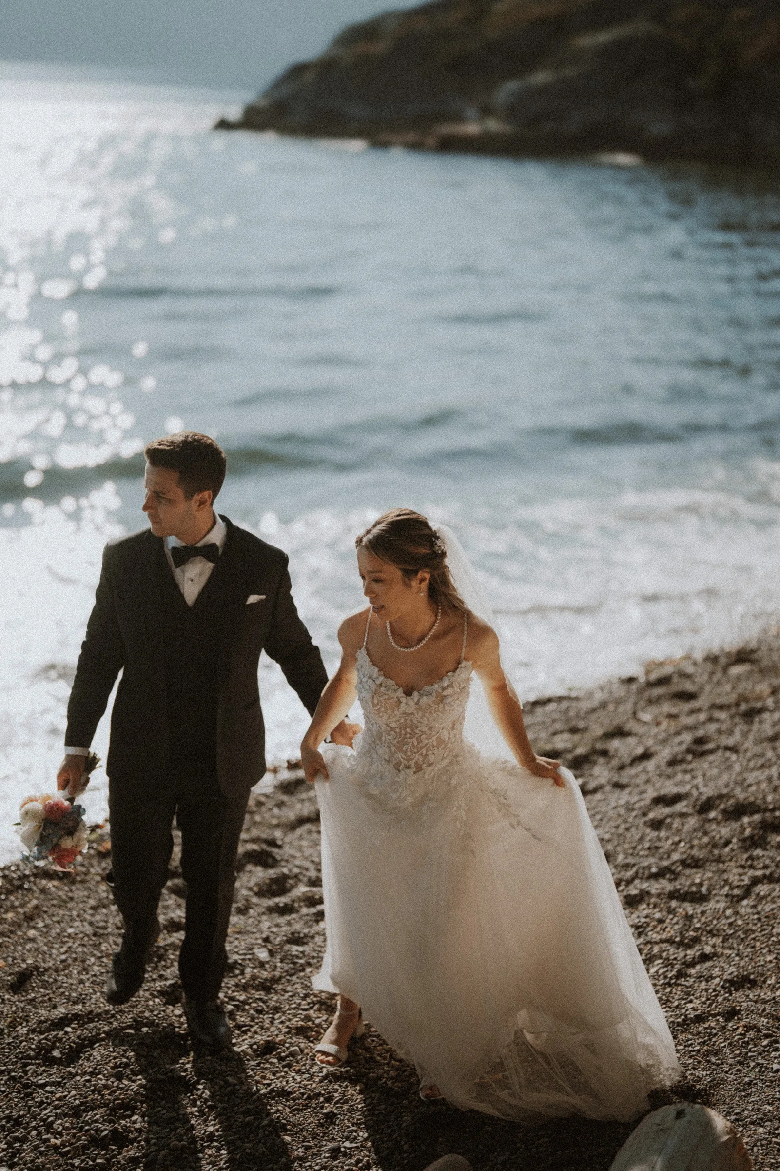 Bride and groom holding hands while walking over pebbles beside the water.