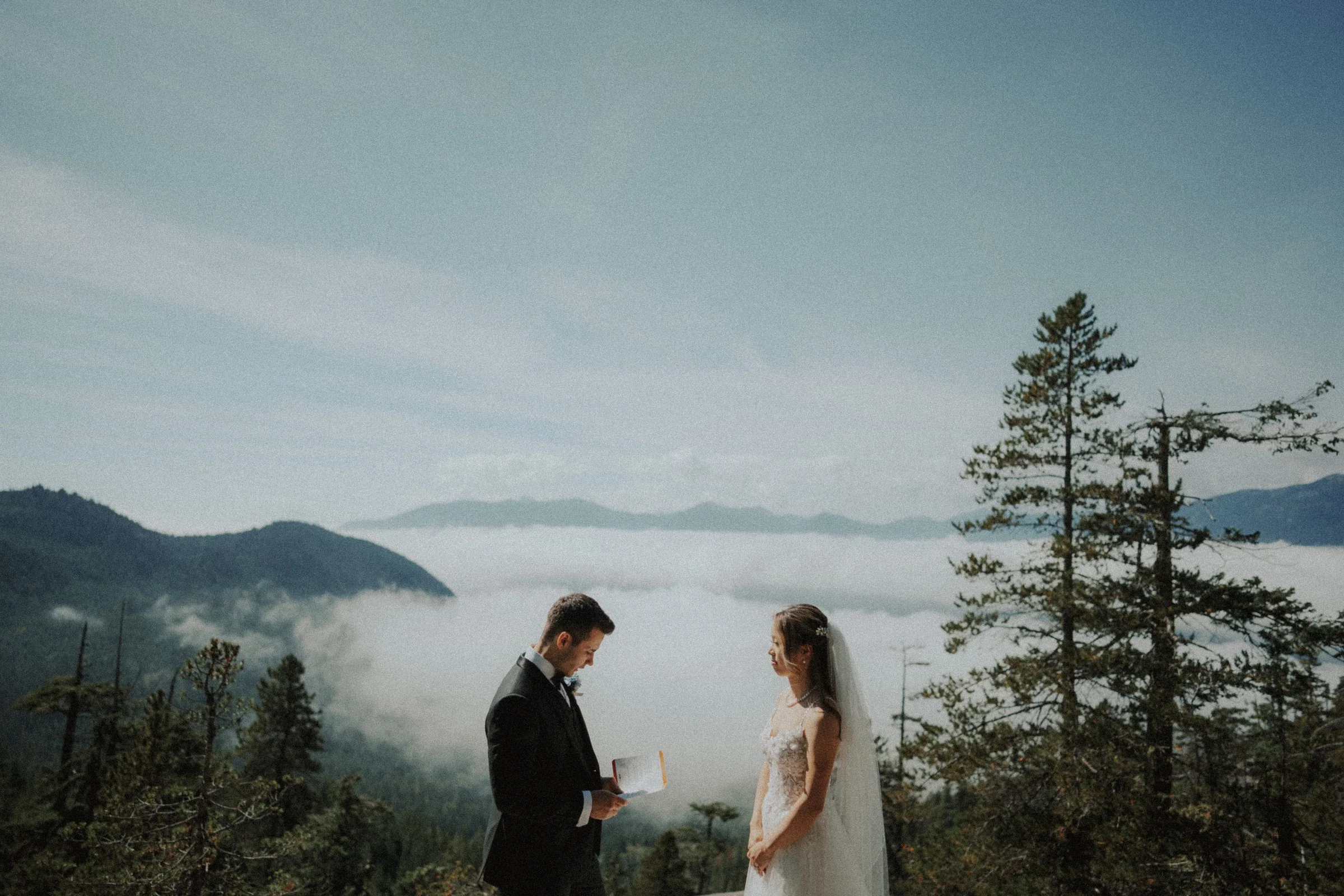 Couple exchanging rings against the mountain and cloud backdrop.