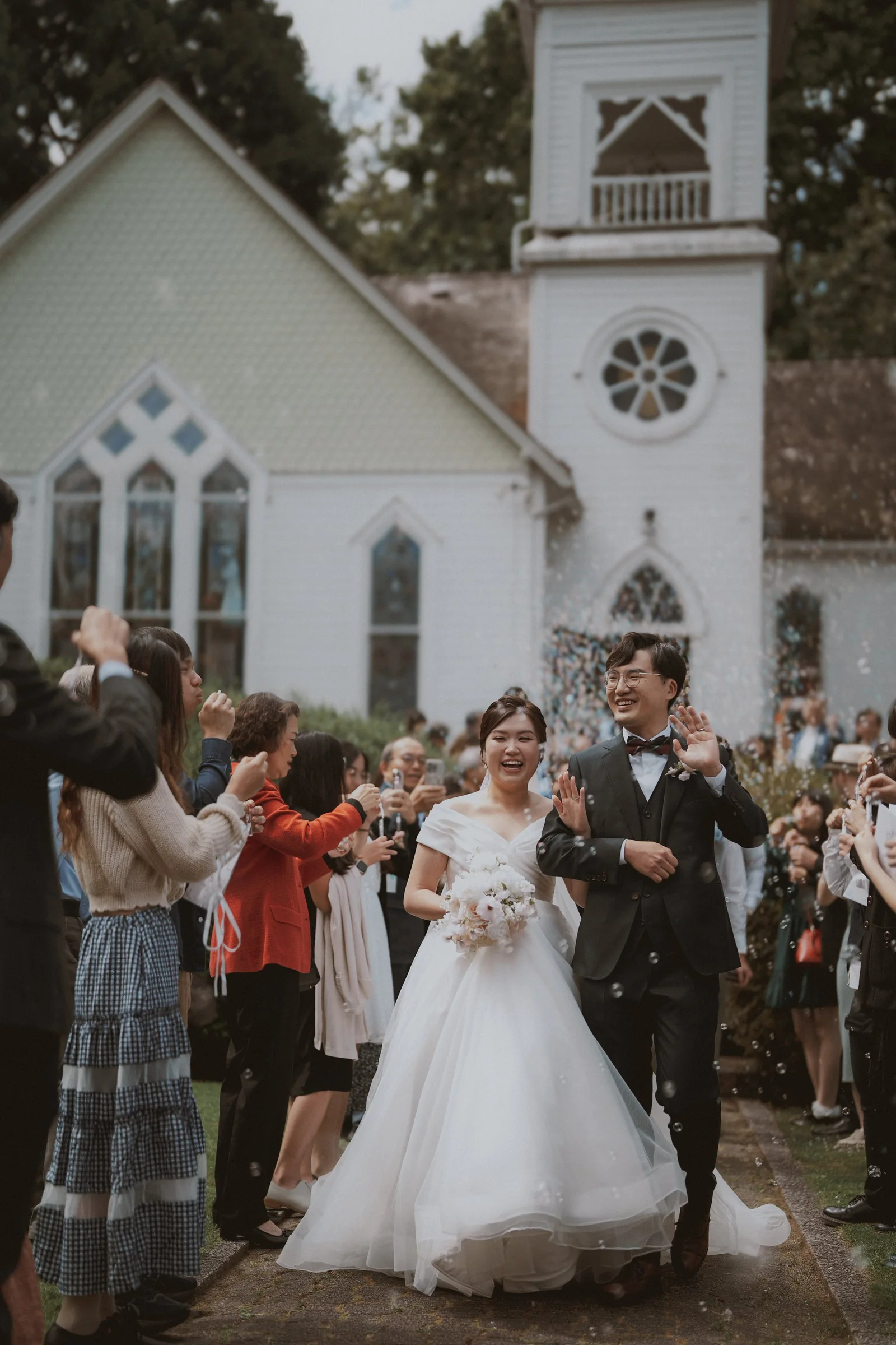 Newlyweds smiling beneath floating bubbles on the chapel garden path.