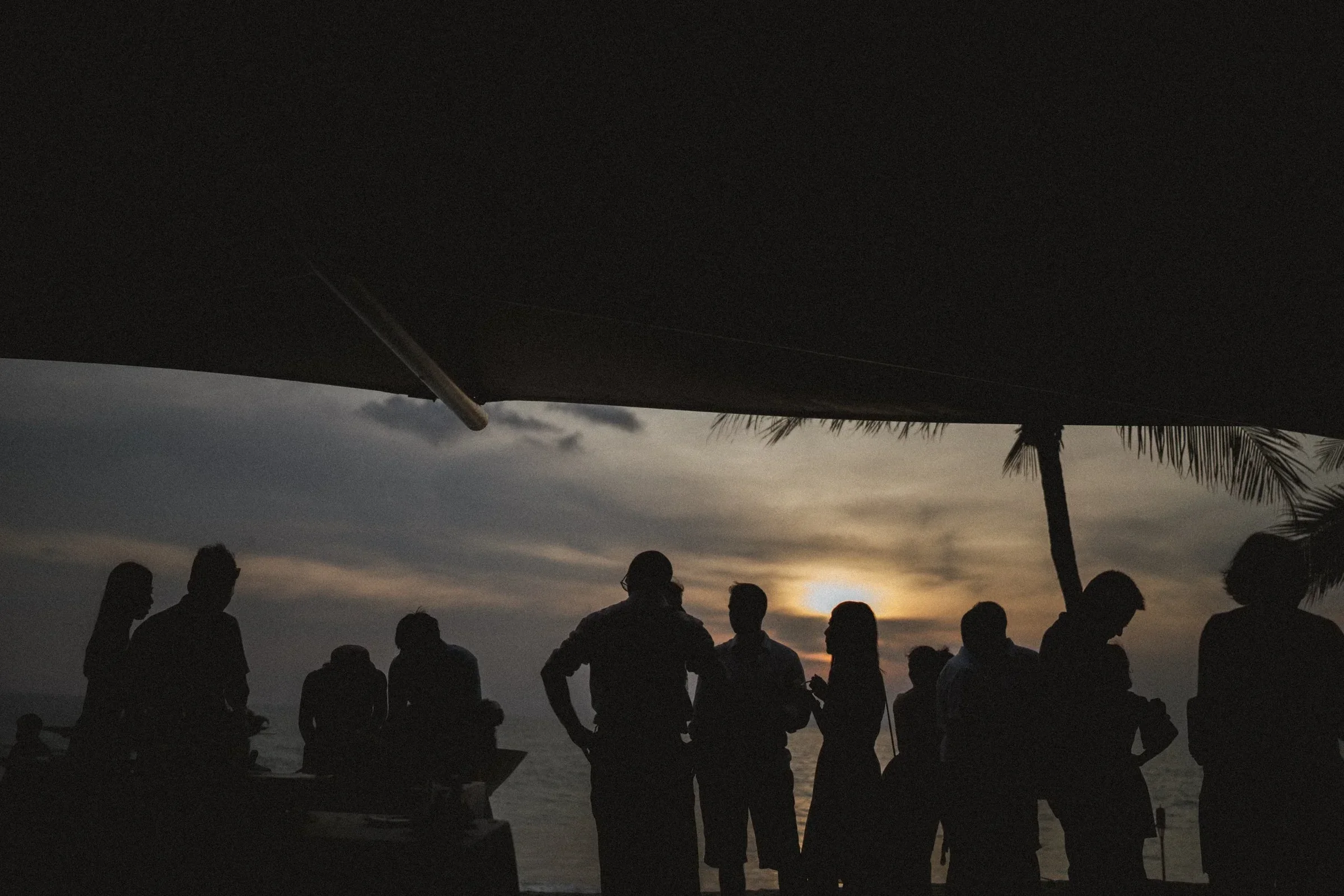 Wedding guests silhouetted against the sea at sunset beneath a covered terrace.