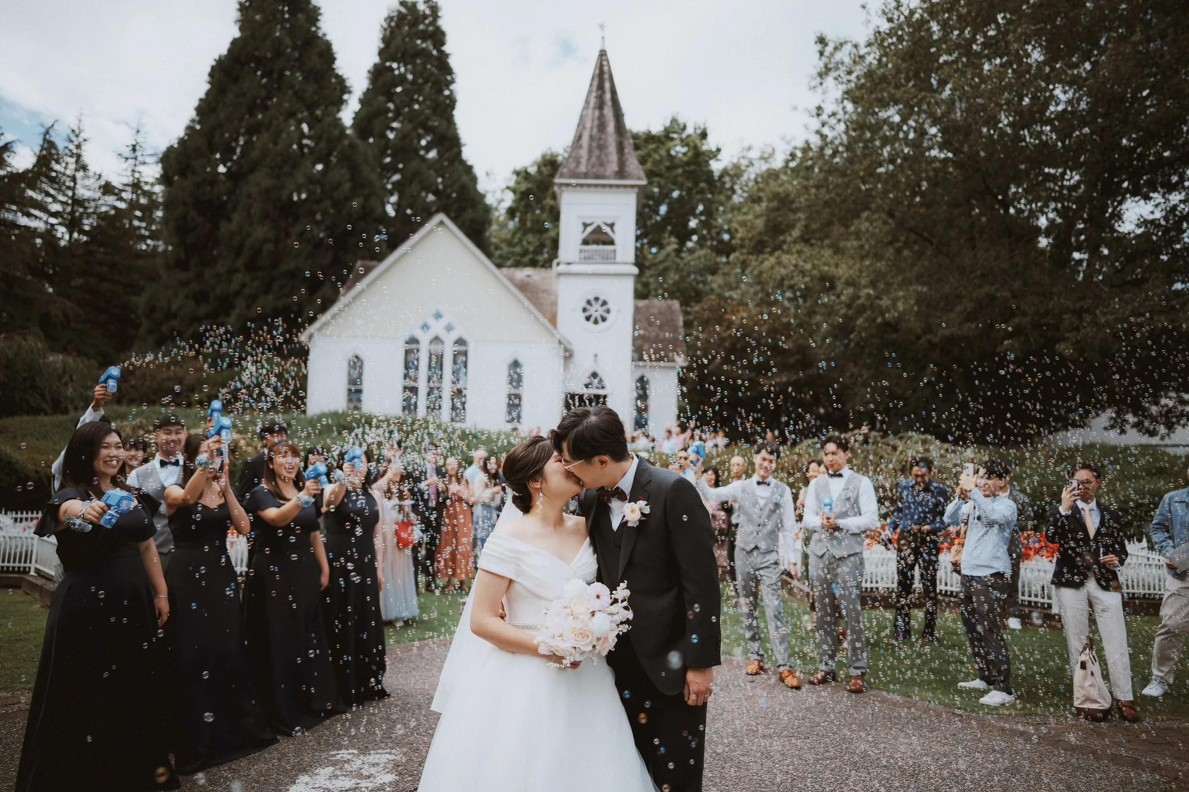 Bride and groom kissing while guests blow bubbles around them.