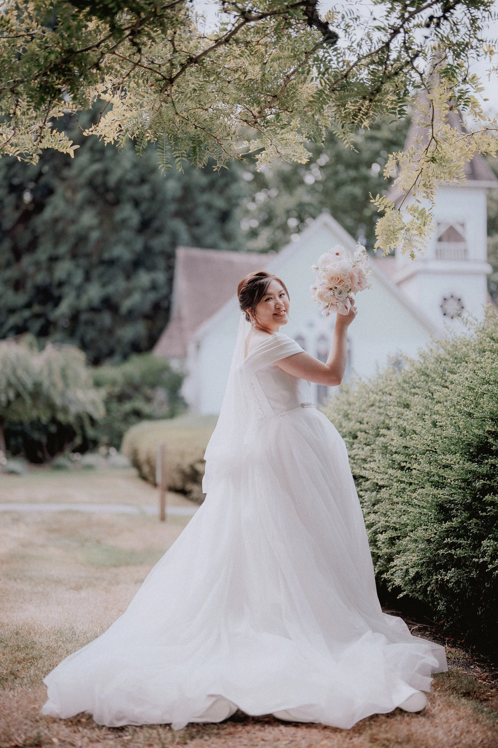 Bride lifting her bouquet beside a hedge with Minoru Chapel behind her.