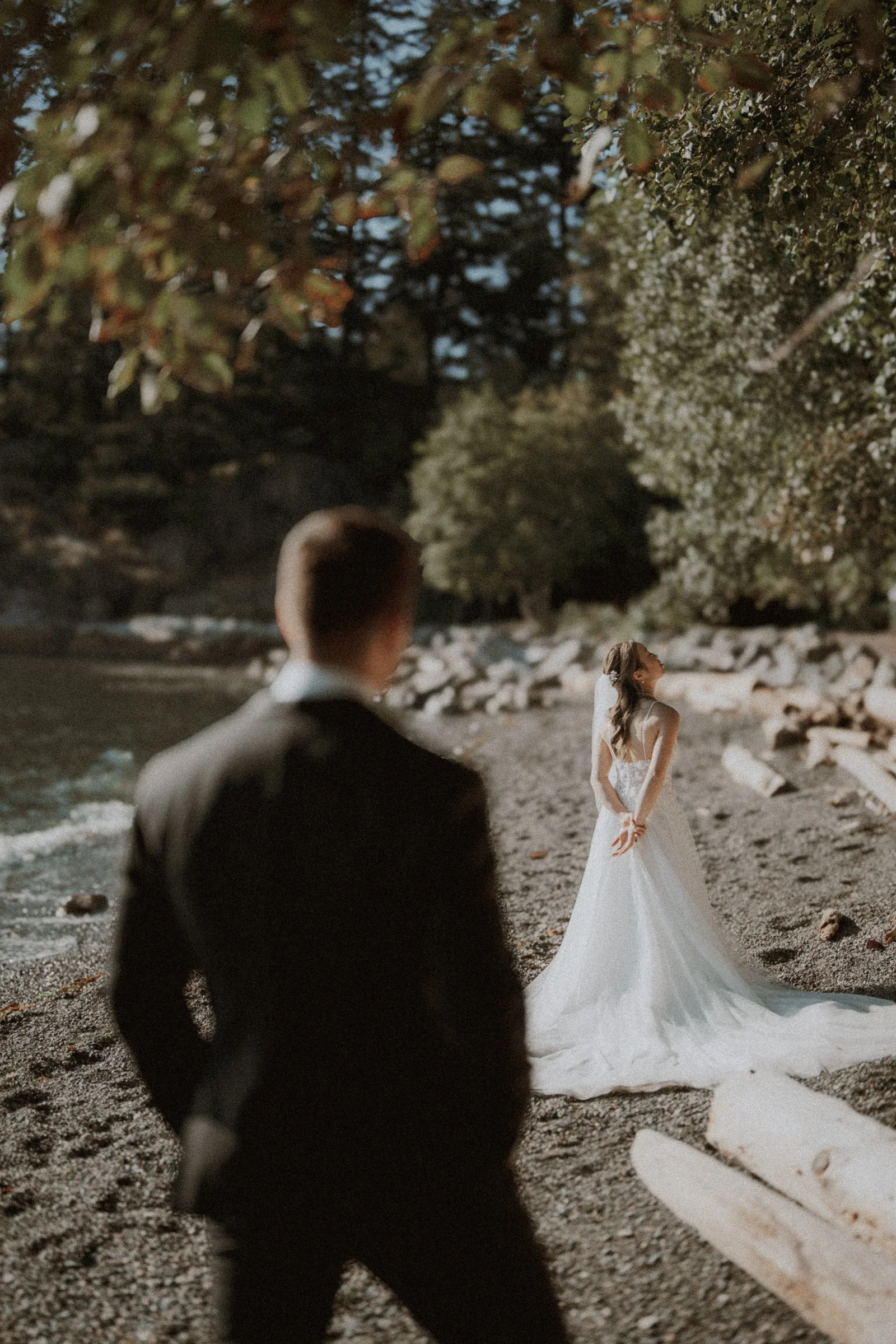 Groom watching the bride near driftwood on the pebbled beach.