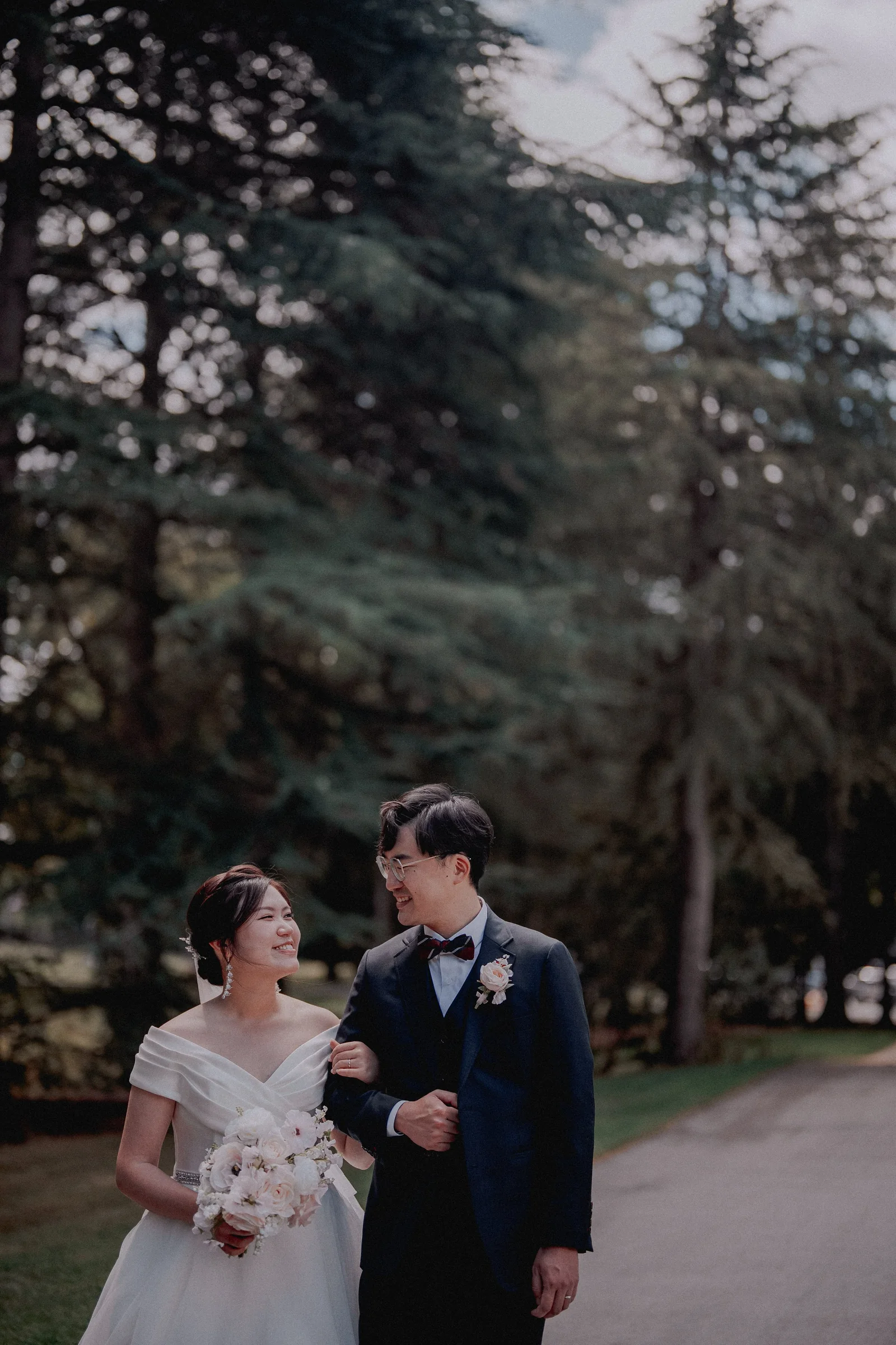 Newlyweds strolling together beneath tall evergreens.