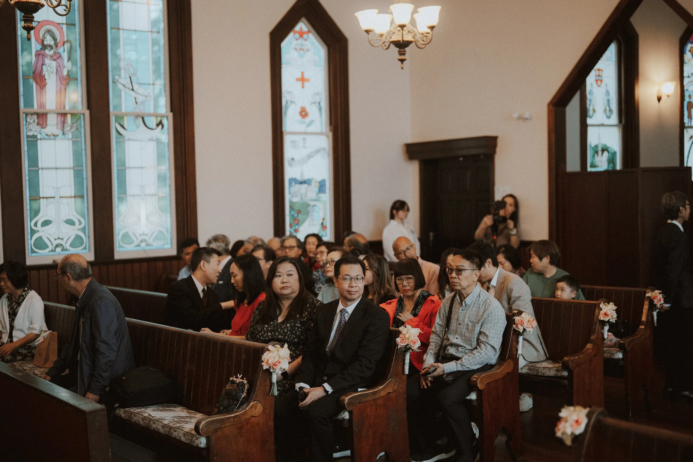 Wedding guests seated in wooden pews beneath stained-glass windows.