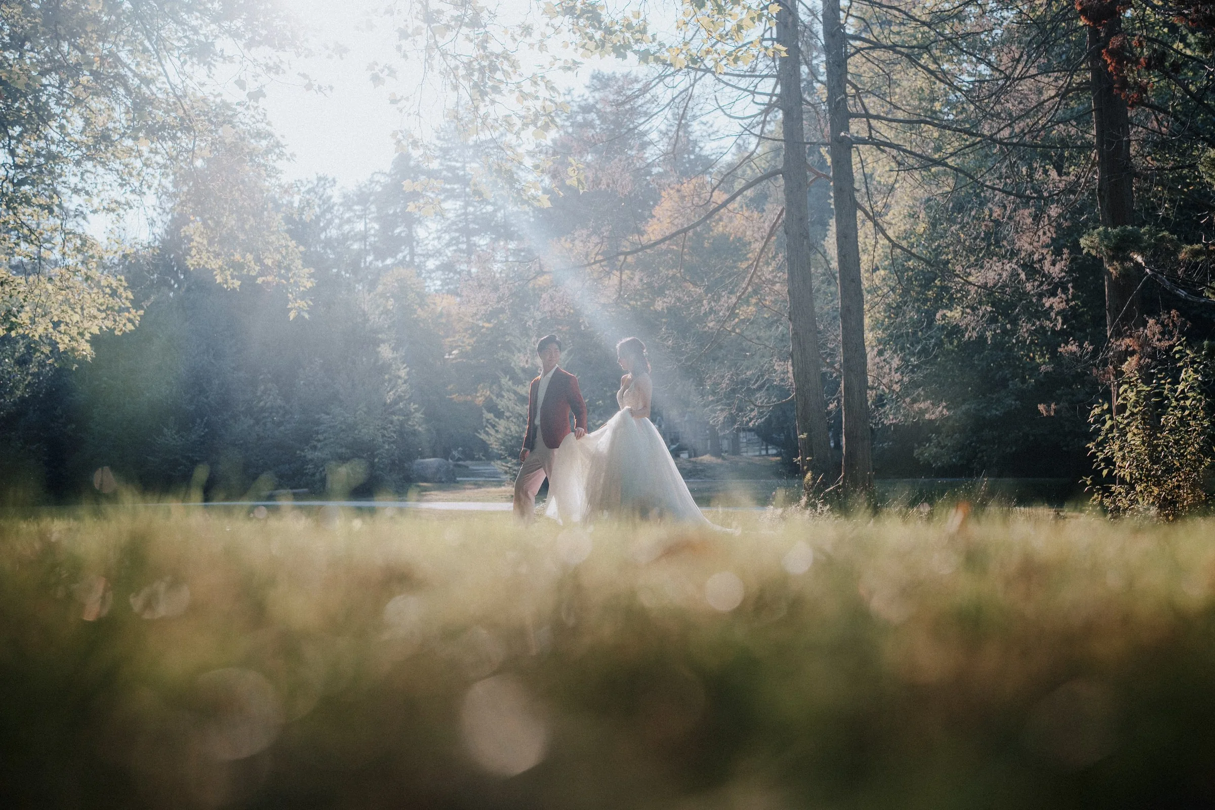A bride and groom standing in a sunlit forest clearing during their wedding photoshoot, with sunlight streaming through the trees