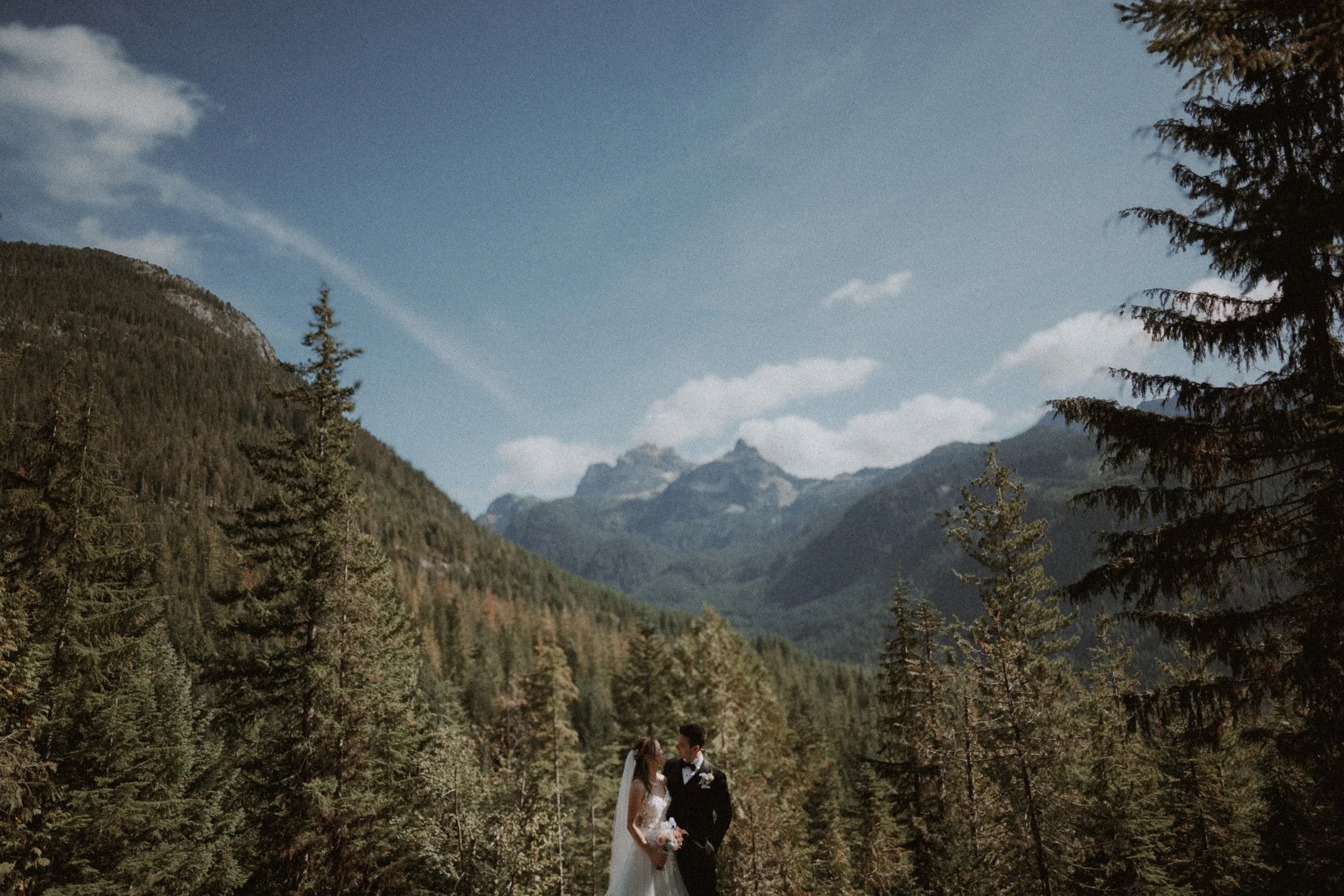 Newlyweds on a mountain trail beneath a wide blue sky and distant peaks.