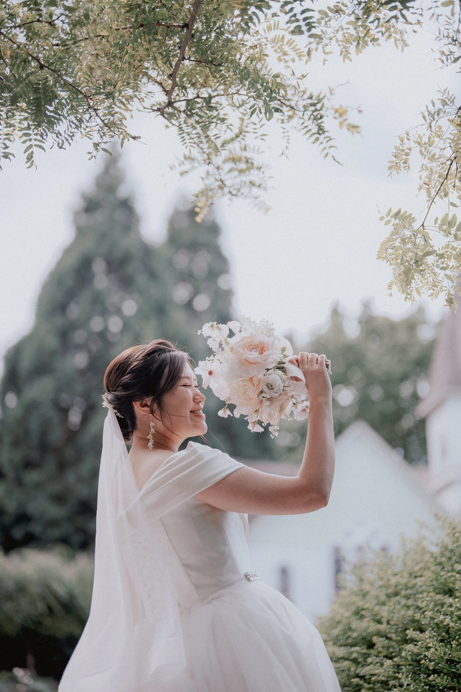 Bride raising her bouquet beneath the branches of a garden tree.