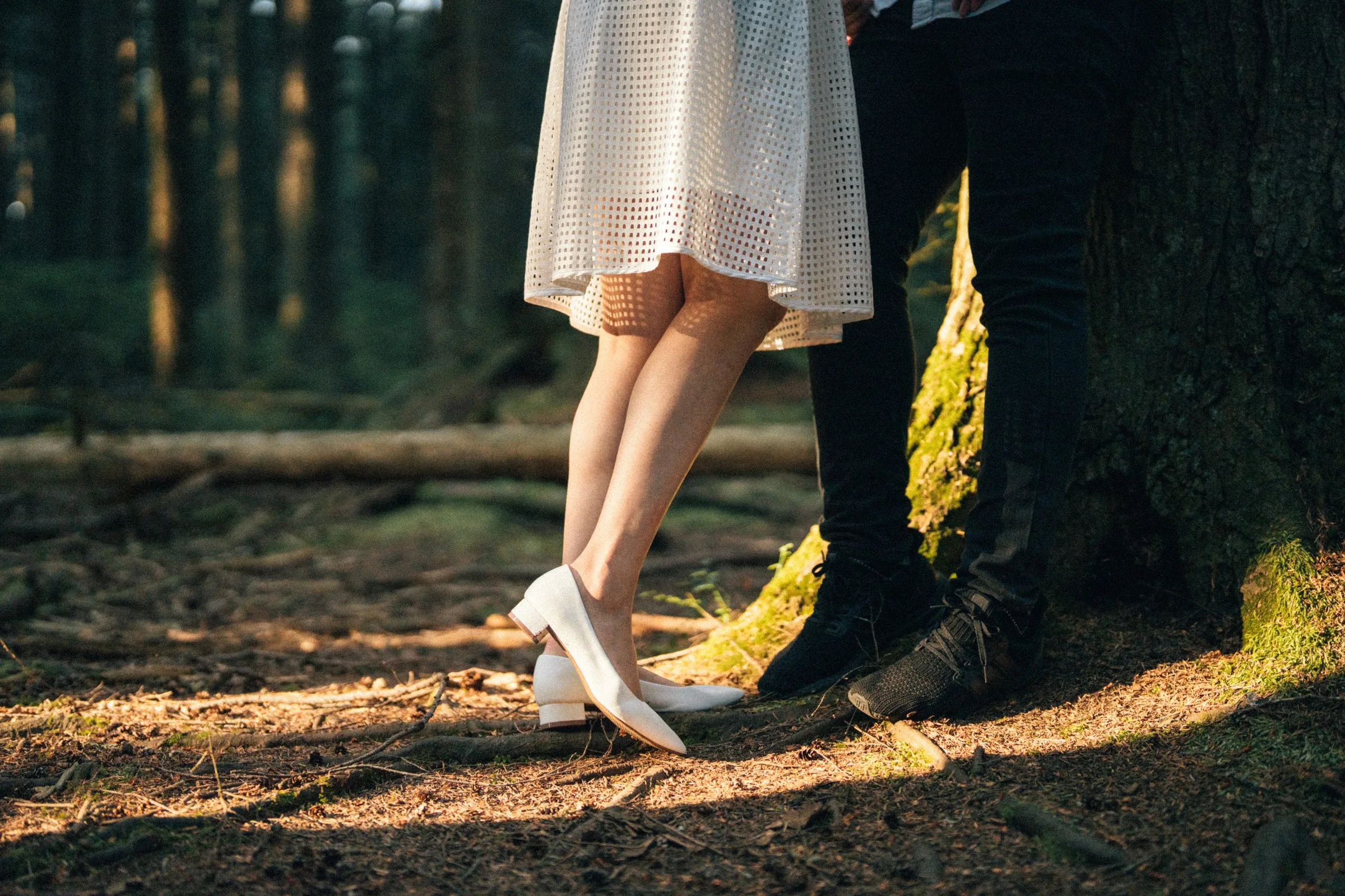 Couple's legs close-up leaning on a mossy tree trunk, warm sunlight.