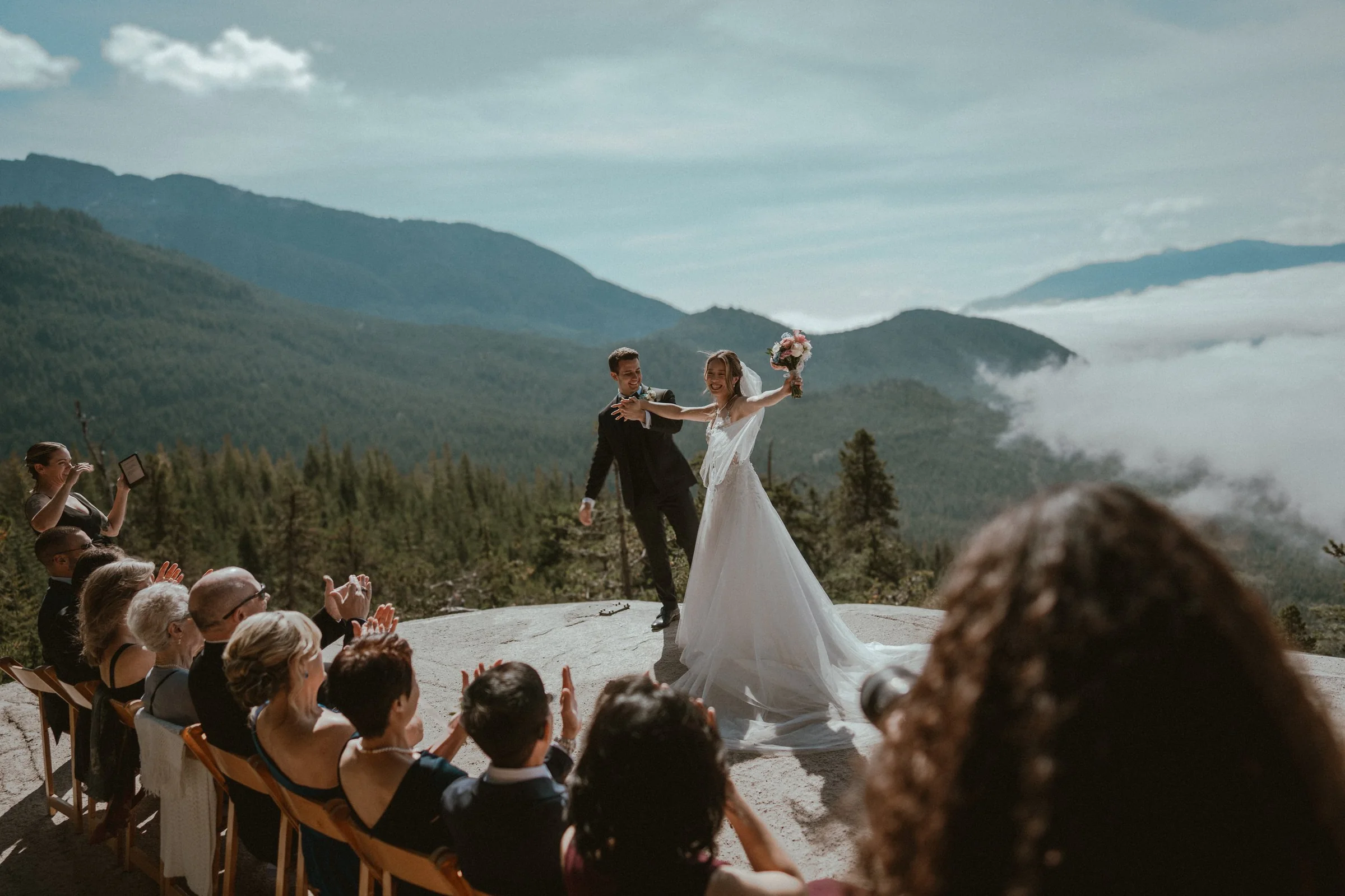 A wedding ceremony on a mountain with a bride and groom, with the bride holding a bouquet and the groom with his hand on her waist, surrounded by seated guests and scenic mountain views in the background.